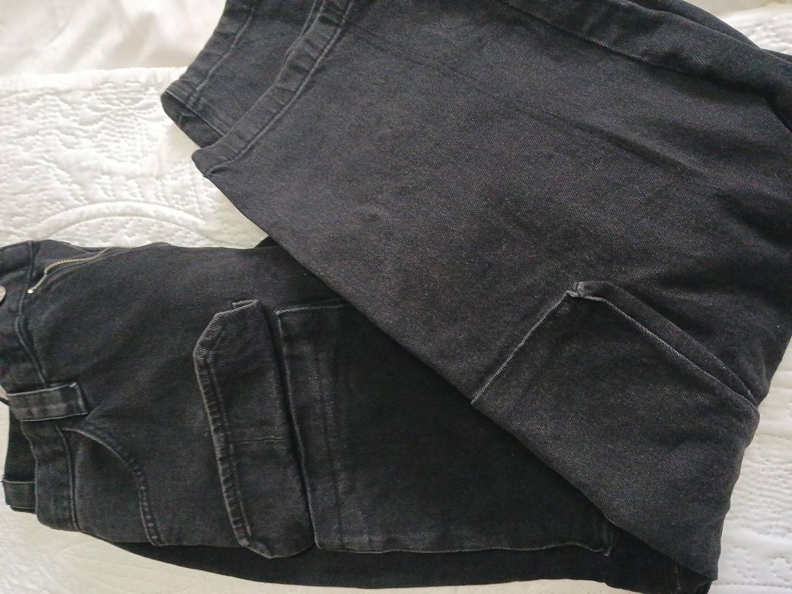 Black denim trousers with pockets, placed on a white quilt with an embroidered design.