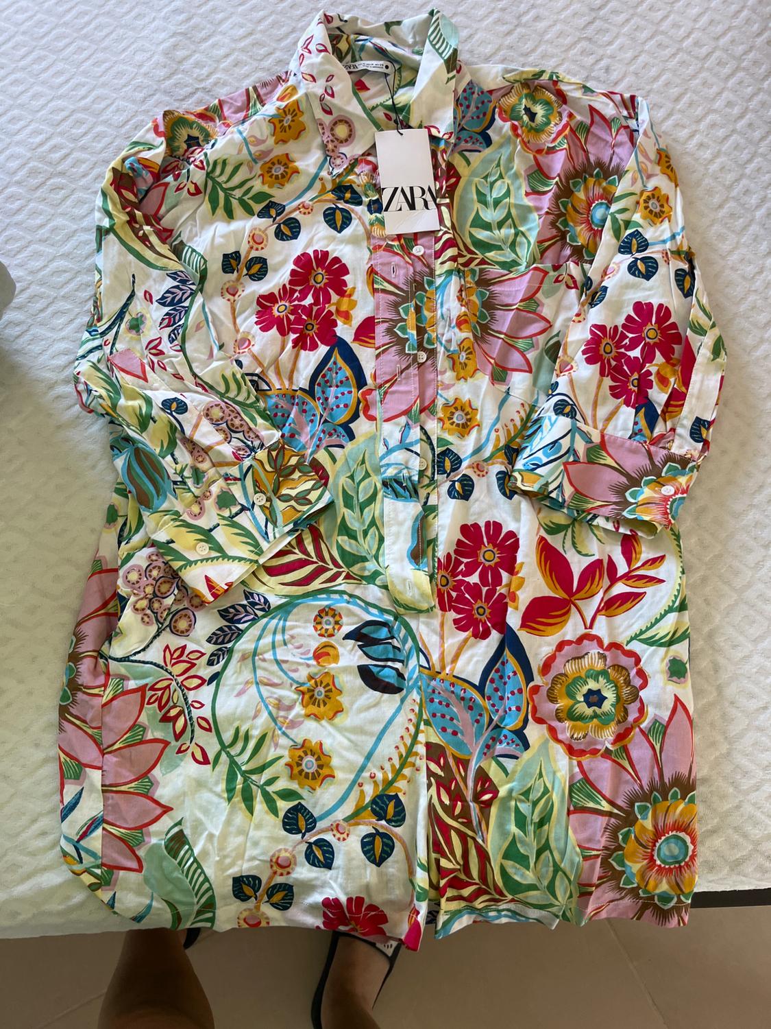 Long shirt with a multicoloured floral print on a white background. Label with ZARA text. Feet in sandals peeking out at the bottom.