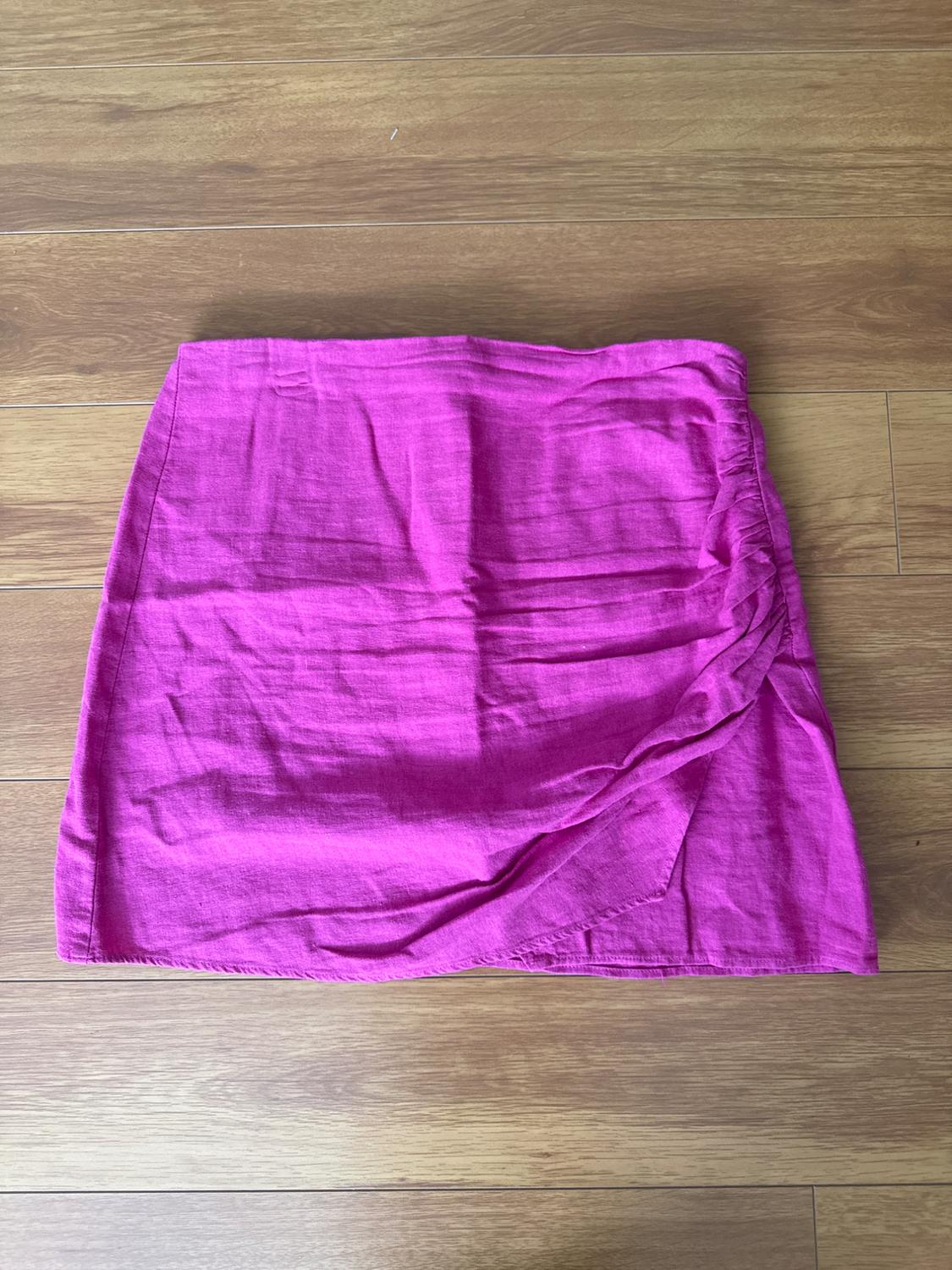 Fuchsia mini-skirt on a wooden floor. Wrinkled fabric with a pleat detail on the right side. No prints or embellishments.