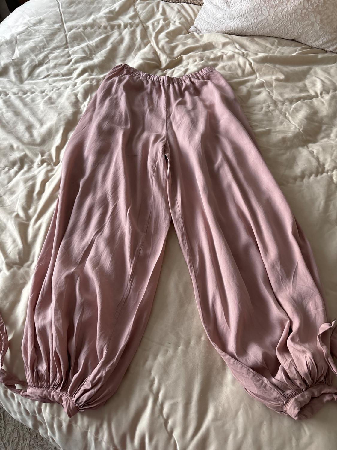 Light pink fluid trousers with an elasticated waistband and gathered hems with ties, laid out on a bed with light-coloured, crumpled-texture sheets.