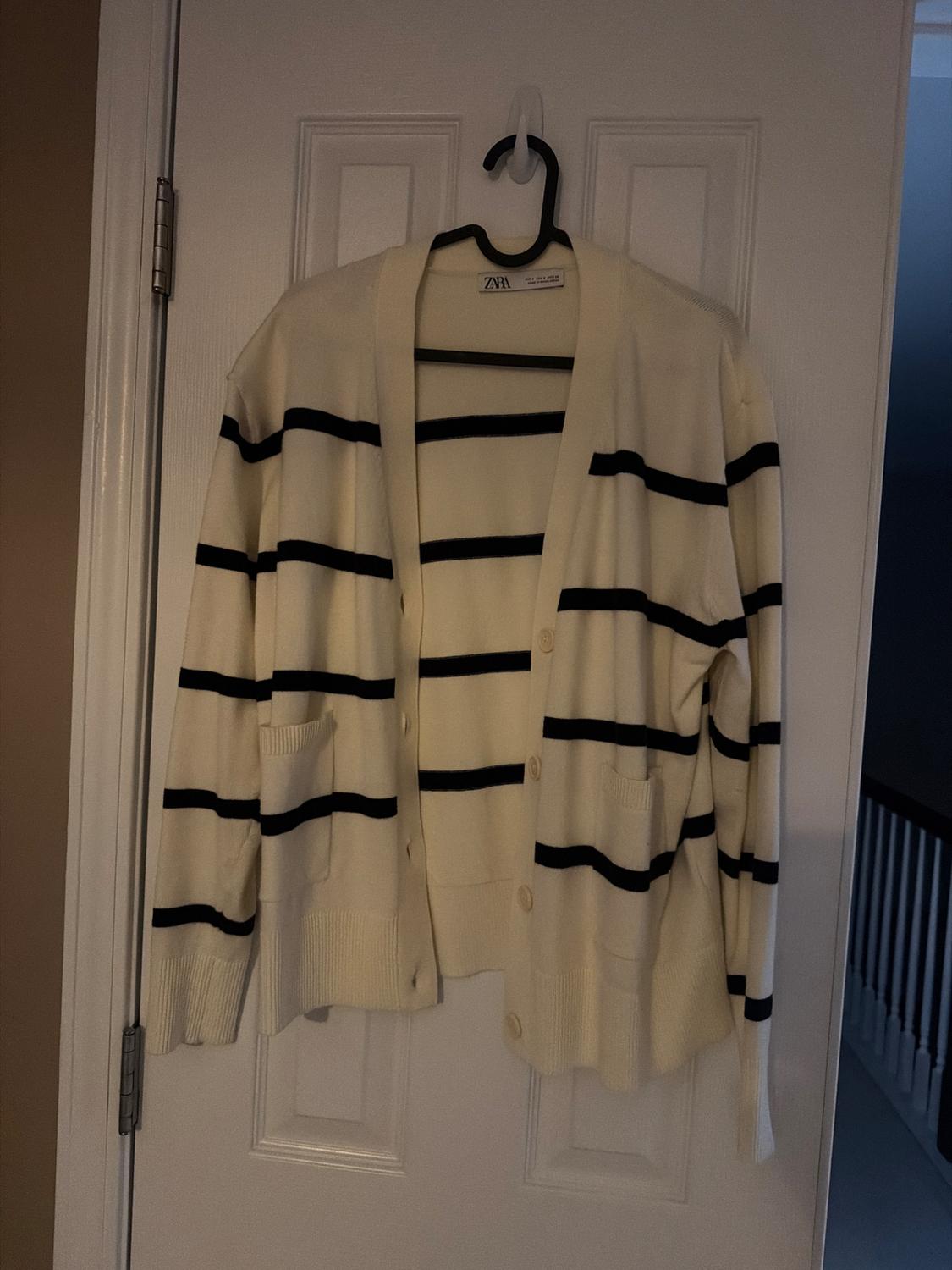 White cardigan with black stripes hanging on a white door. Visible tag with the brand. Indoor setting with dim light.
