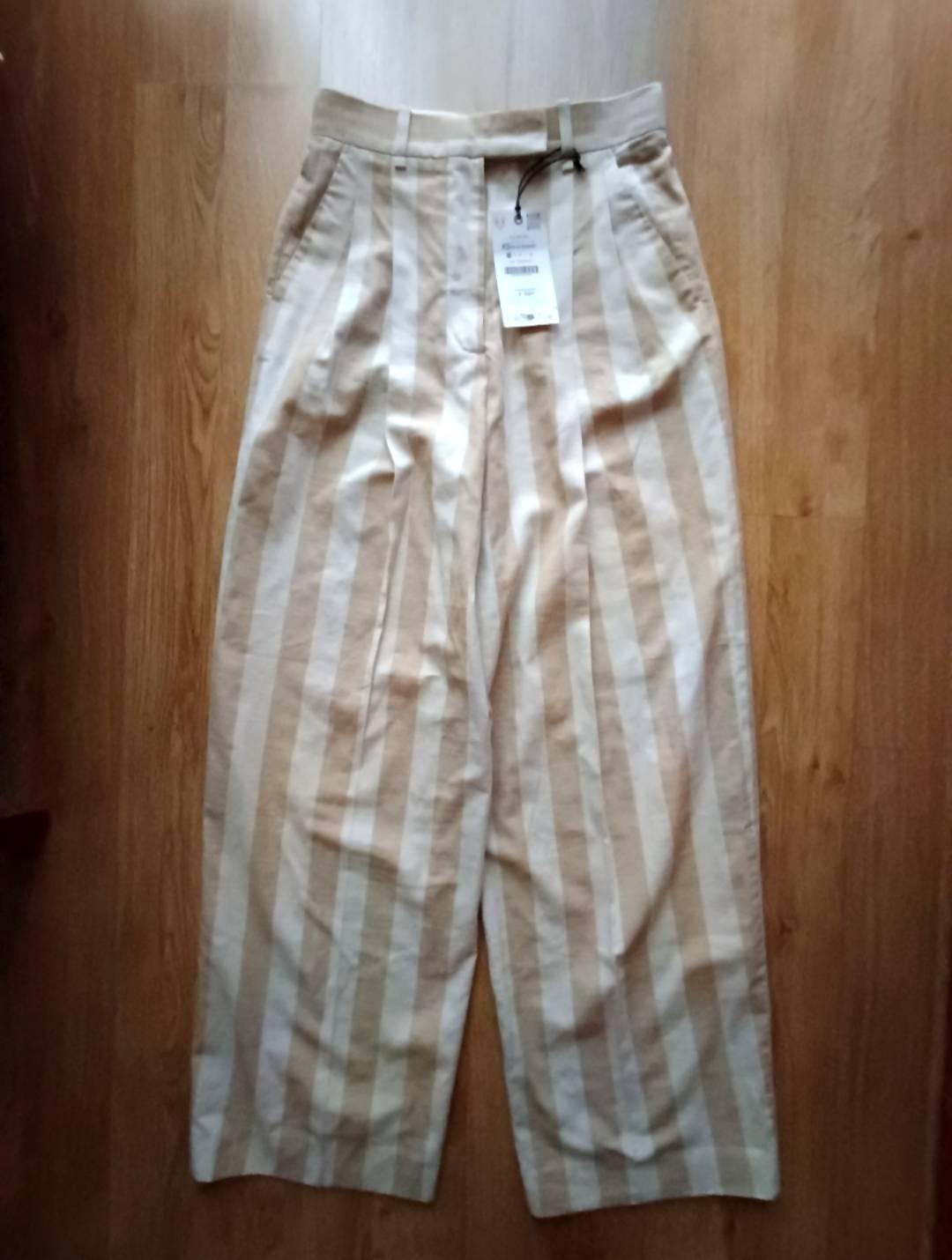 Beige and white vertical striped wide-leg trousers, with pleats and a label, on a wooden floor.