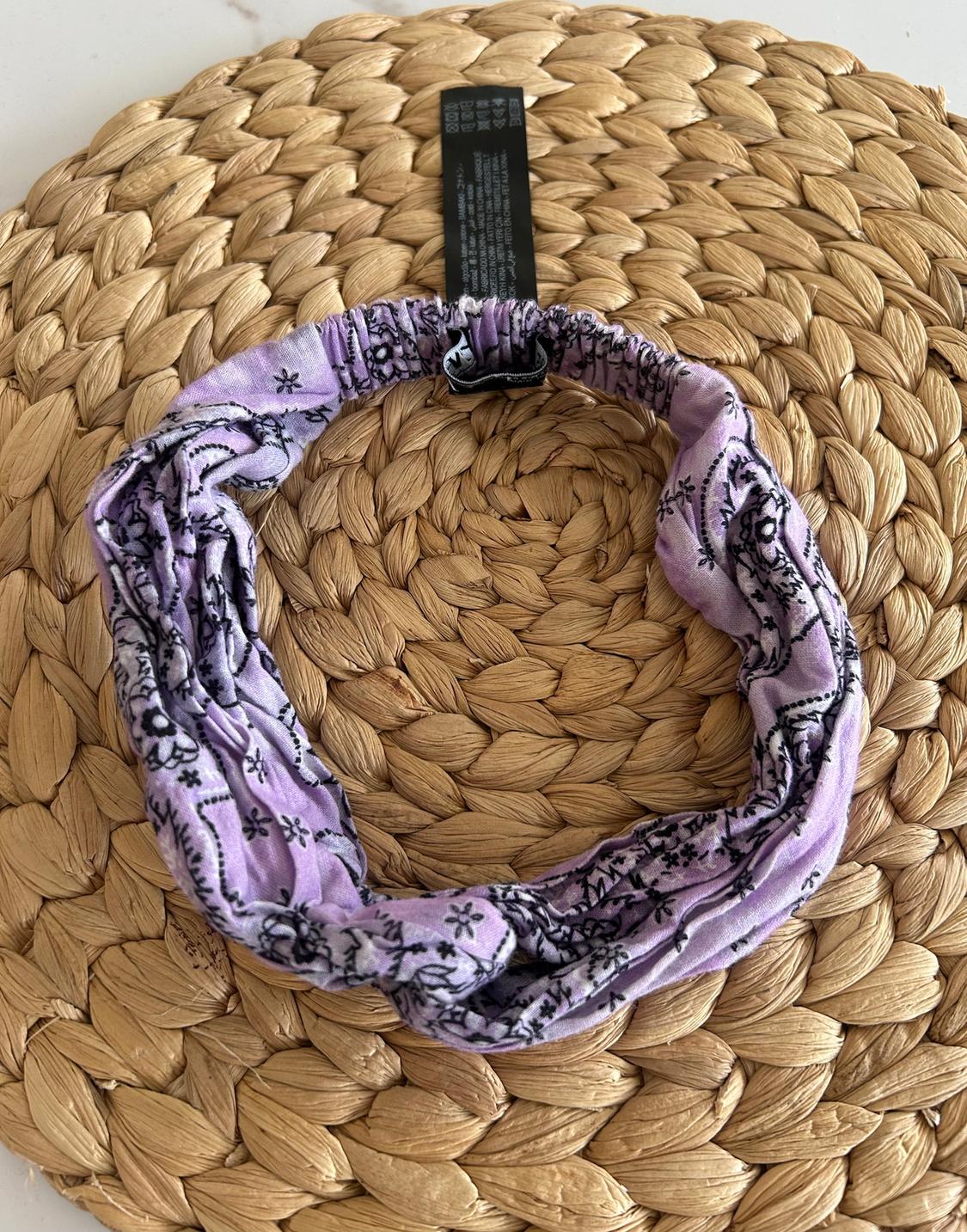 Lilac fabric hairband with black floral print on a braided wicker background. Visible black label.