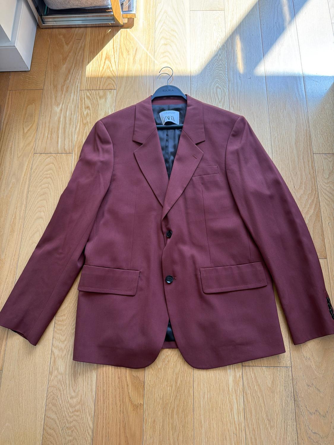Burgundy suit jacket hanging on a black hanger over a light wood floor, with natural light illuminating the scene.