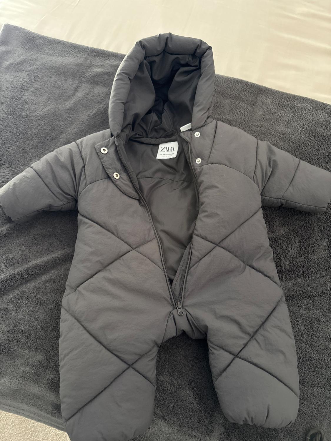 Dark grey padded jumpsuit with hood, zip and button fastening, visible Zara label, on a grey blanket on a beige surface.