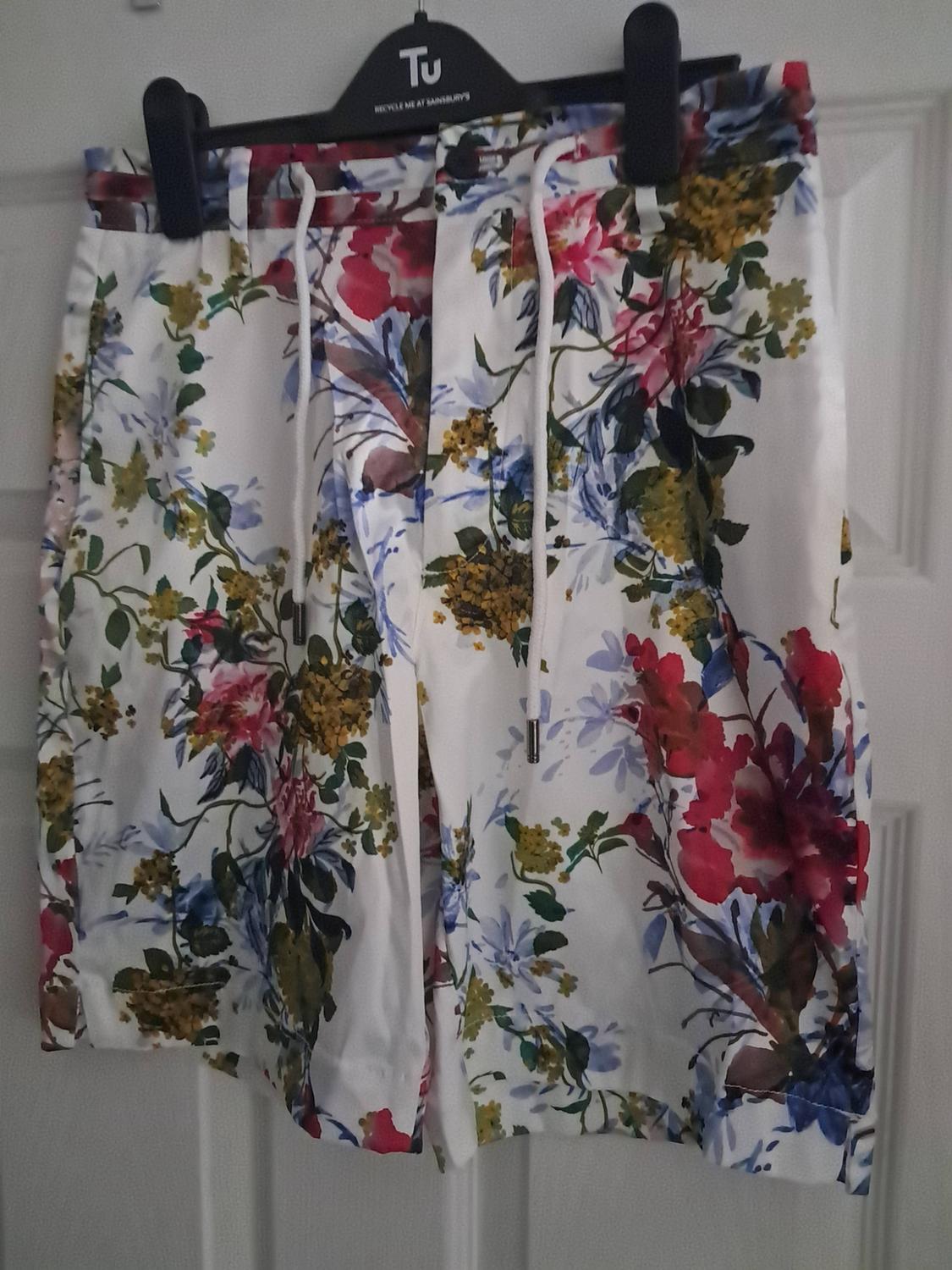 White shorts with a floral print and drawstring, hanging on a door. Text: RECYCLE ME AT SAINSBURY'S.