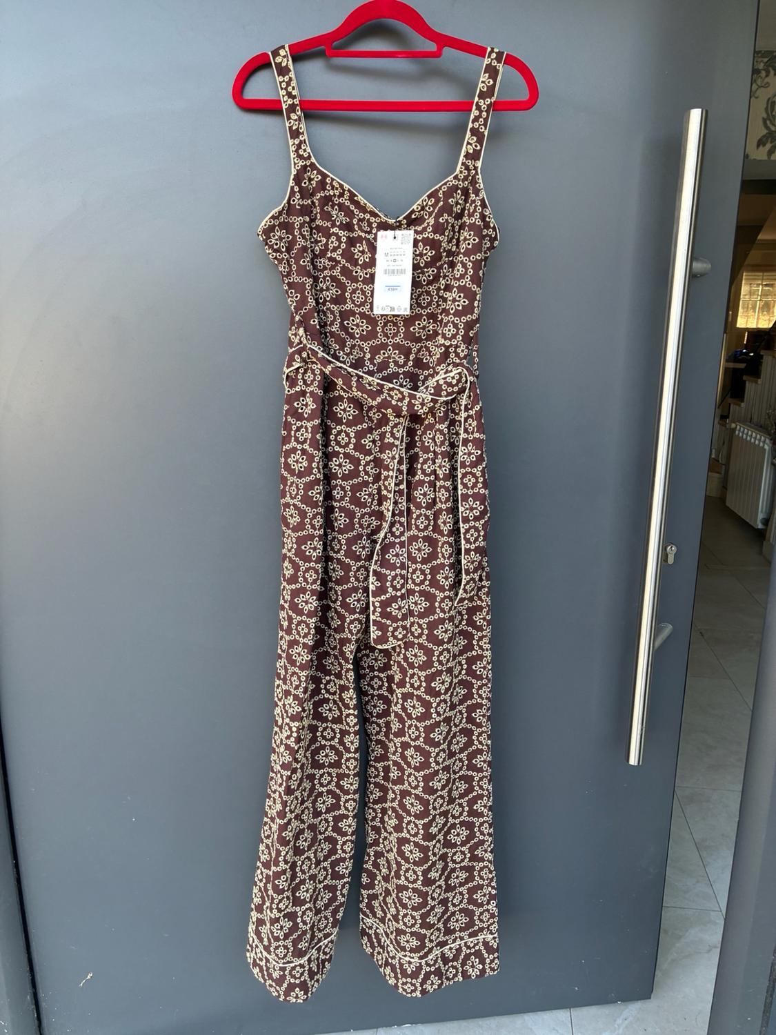 Long brown jumpsuit with a white pattern, V-neckline, shoulder straps and a belt. It is hanging on a red hanger on a grey door.