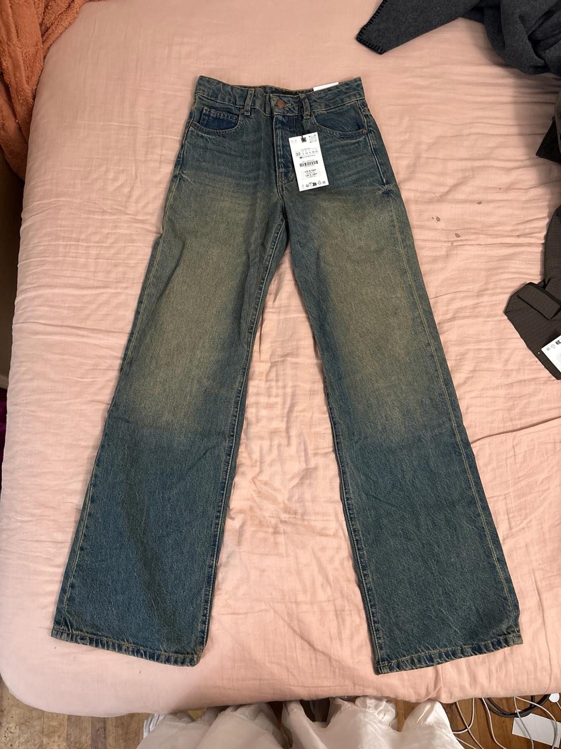 Blue straight-leg denim jeans with a distressed wash and label, laid out on a bed with pink sheets.