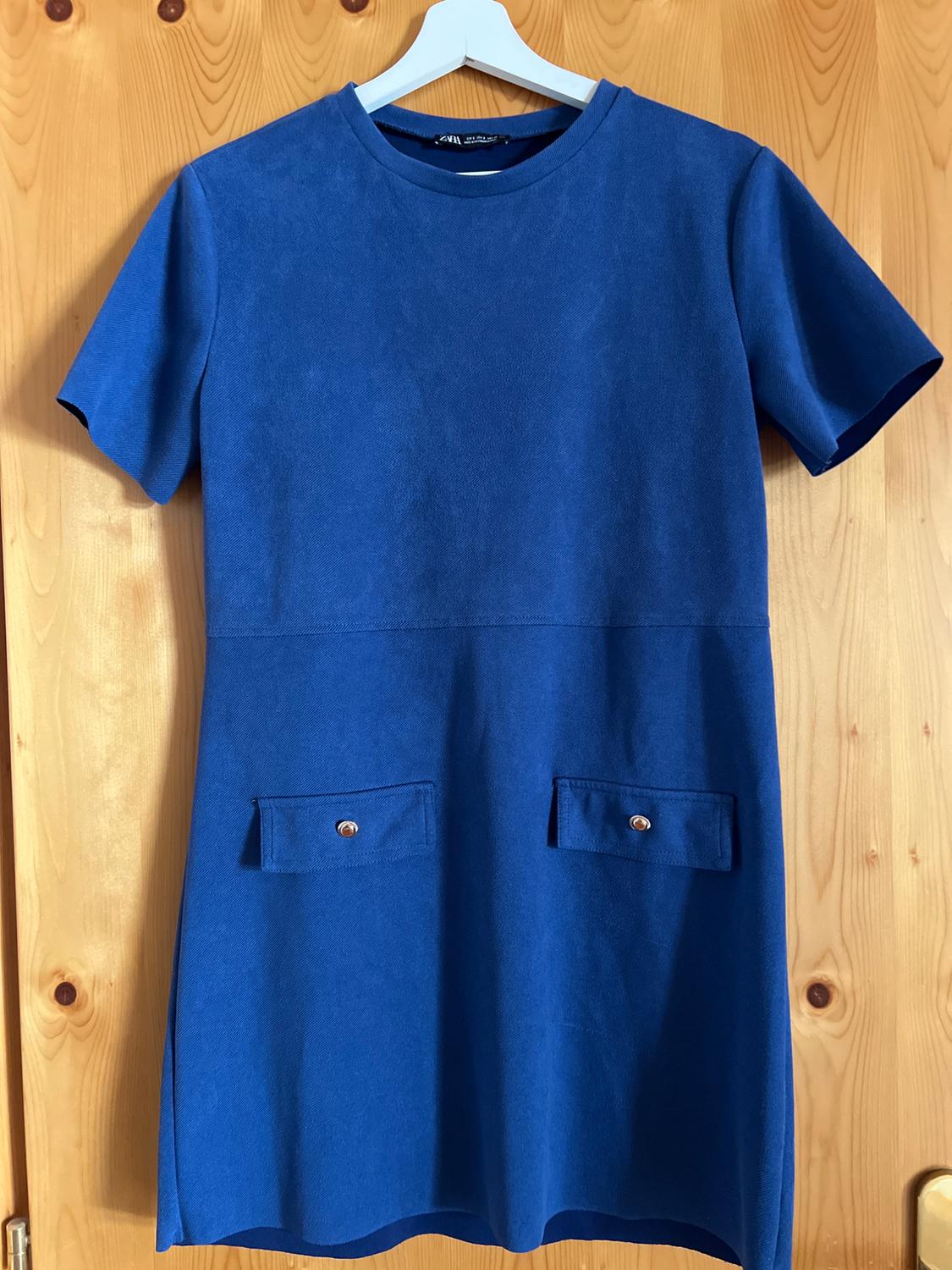 Short-sleeved blue dress with a round neckline and pockets with metallic buttons. ZARA label. Hanging on a hanger on a wooden door.