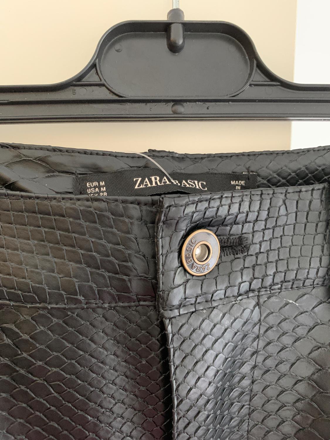Faux leather trousers with a scale texture, metal button and "ZARA BASIC" label on the waistband, hanging on a black hanger.