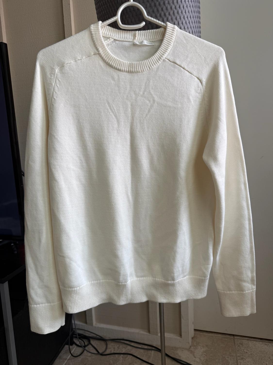White long-sleeve knit sweater with a crew neck, hanging on a hanger in a casual home setting.