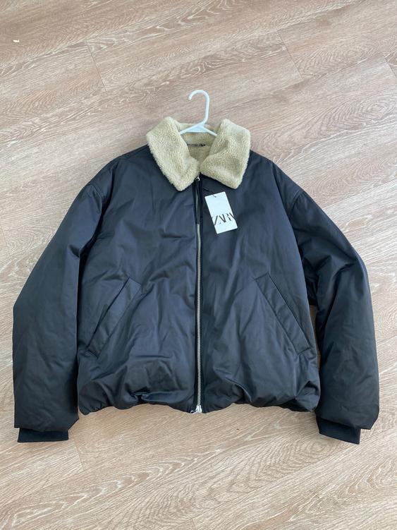 Black jacket with a zipper and a beige shearling collar, hanging on a white hanger over a light wood floor. "ZARA" tag.