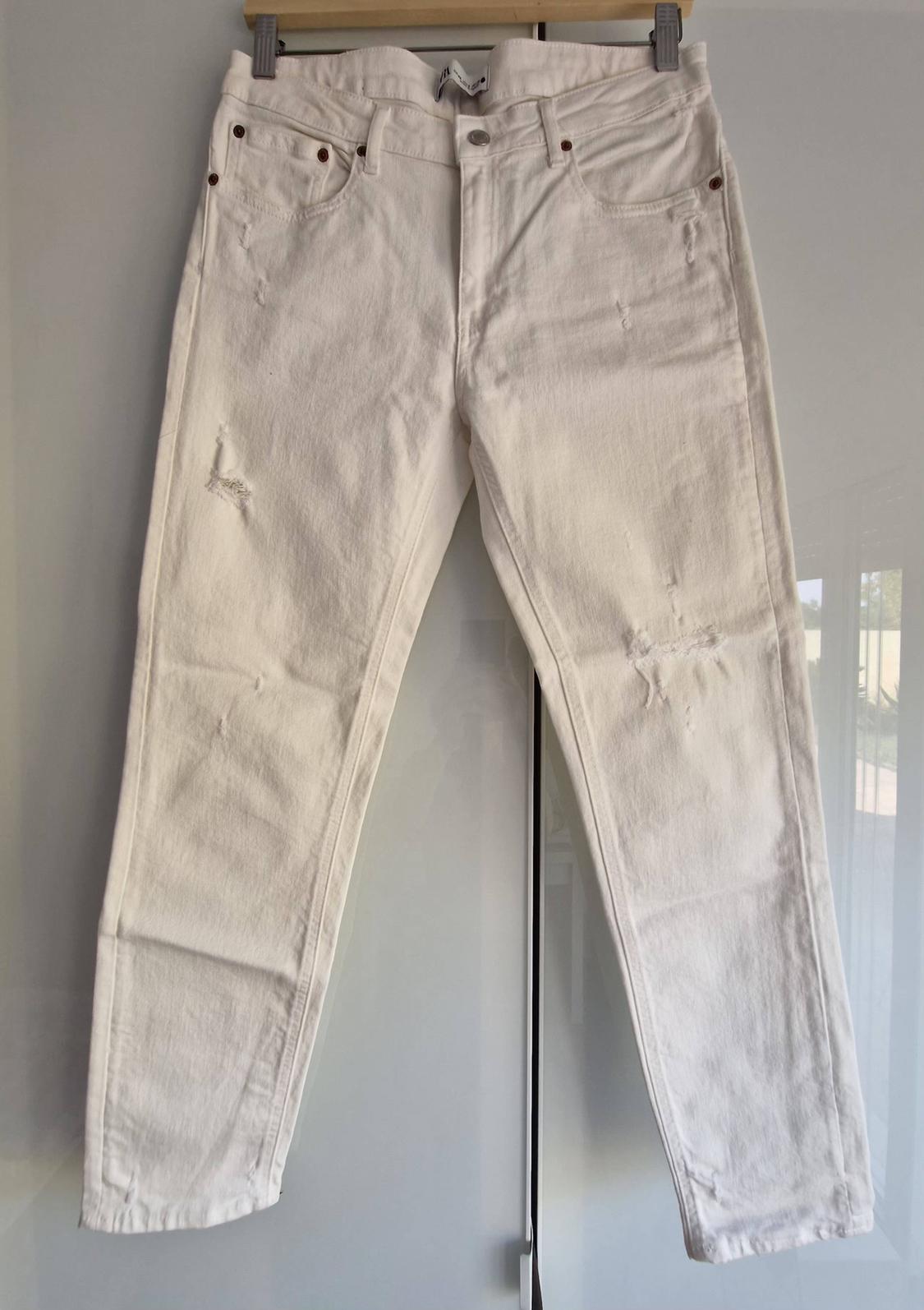 White ripped denim trousers hanging from a hanger above a glossy white surface with reflections.