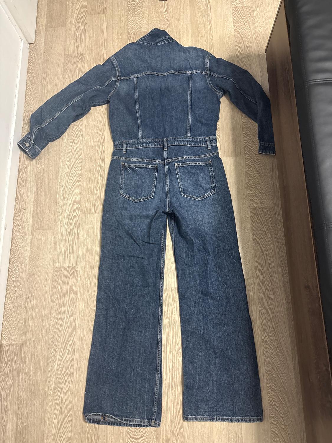 Blue denim one-piece jumpsuit, with back pockets, spread out on a light wooden floor.