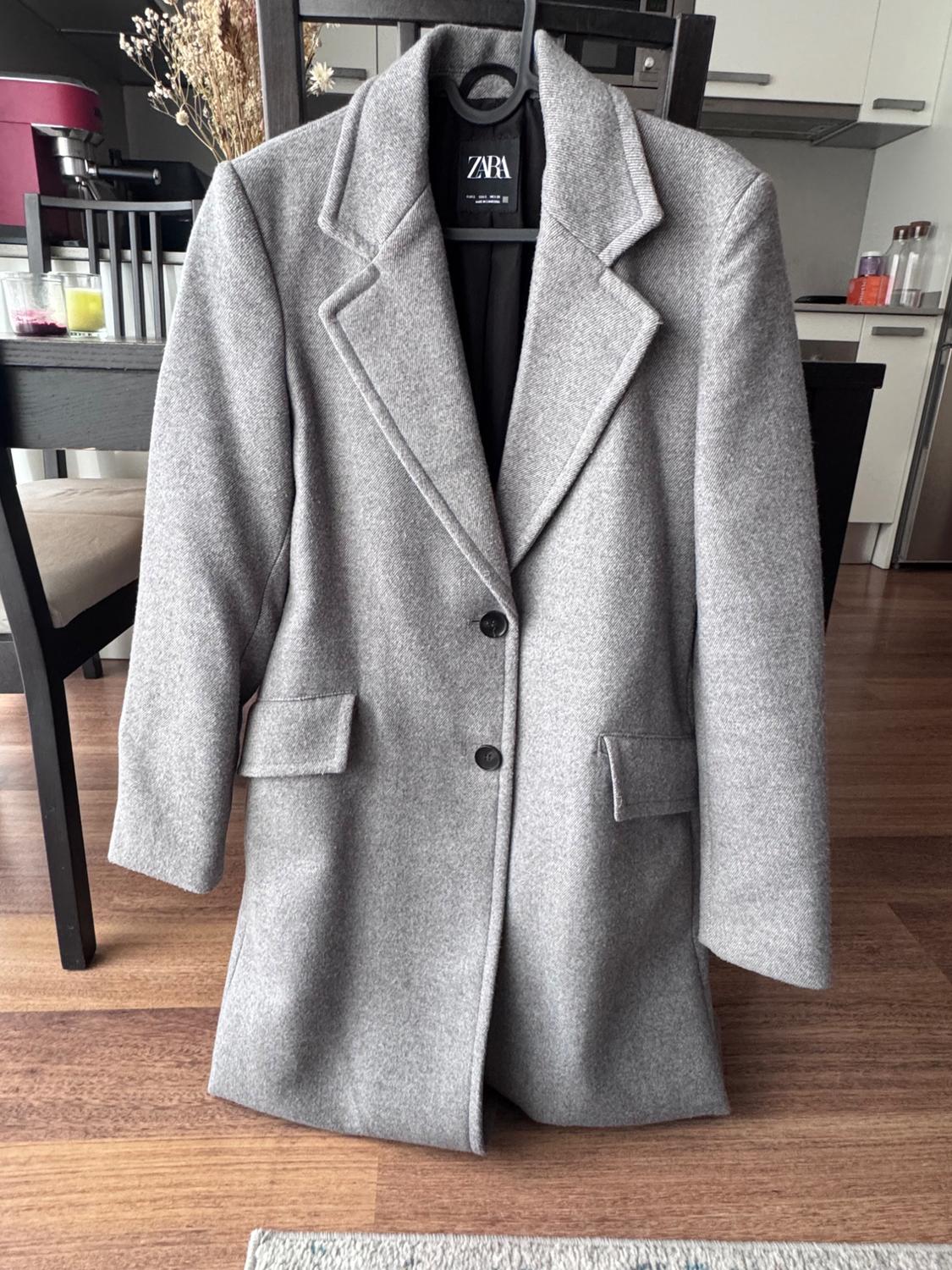 Grey wool coat hanging on a hanger, with two buttons and wide lapels. Dining room background with wooden table and chairs.