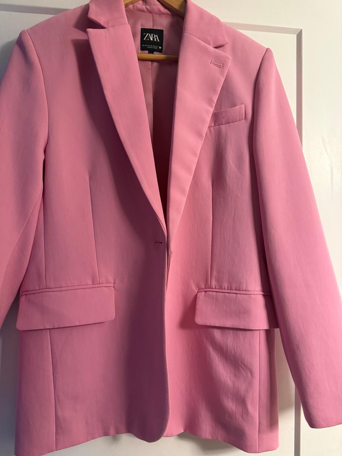Pink jacket hanging on a hanger, with classic lapels and flap pockets. Visible label with the text "ZARA".