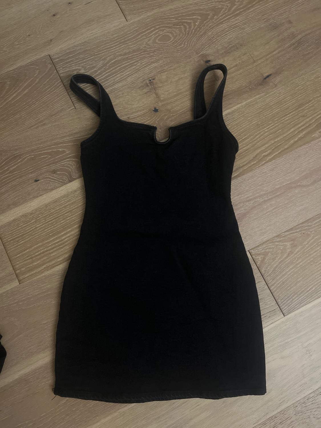 Sleeveless black dress on light wooden floor, with wide straps and a round neckline.