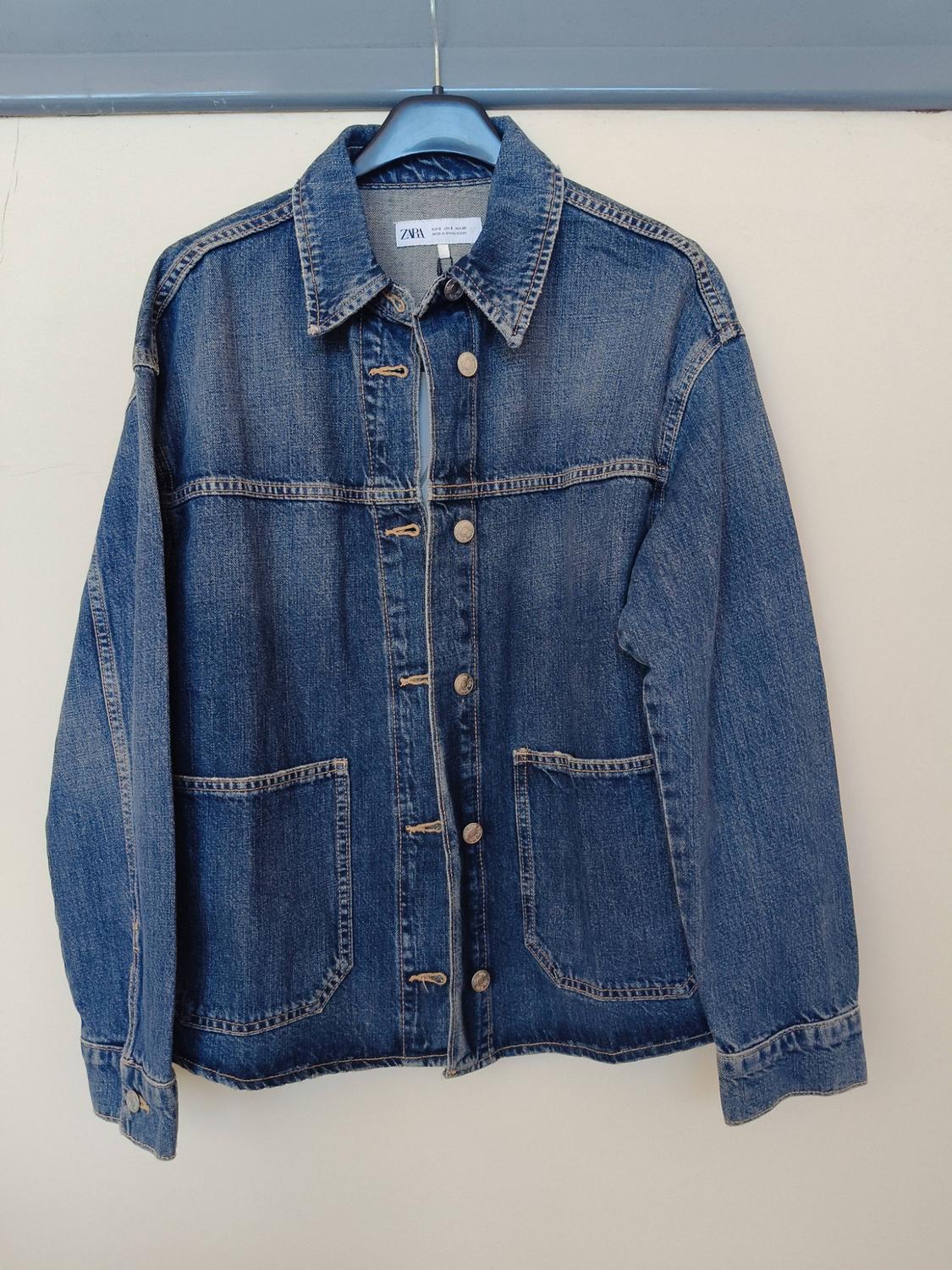 Blue denim jacket hanging on a hanger against a light-coloured wall. It has buttons, front pockets, and a label with the text ZARA.