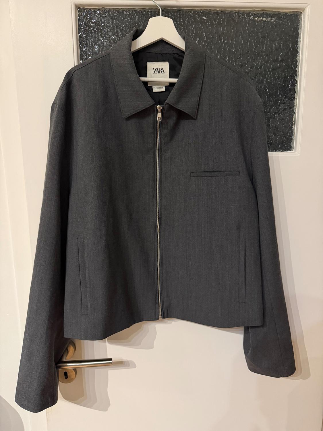 Grey jacket with zip and classic collar hanging on a white hanger. Visible label with the text "ZARA". White door in the background.
