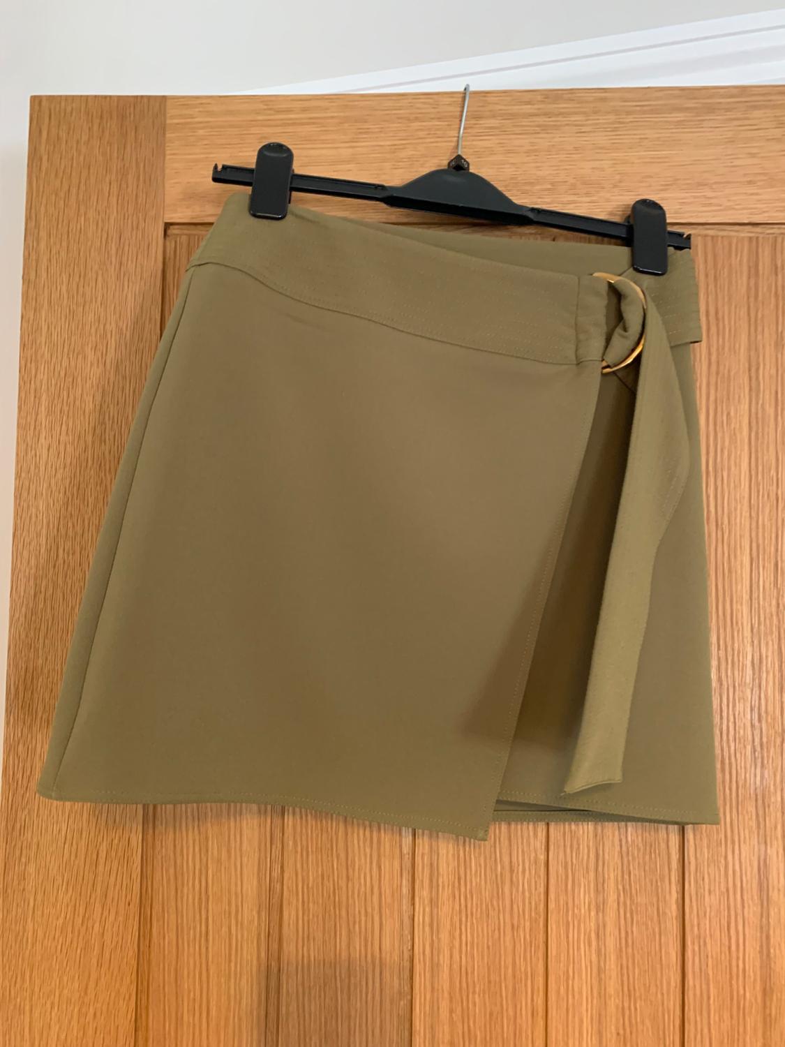 Khaki-coloured mini skirt hanging on a black hanger above a wooden door. Simple design with a knotted belt.