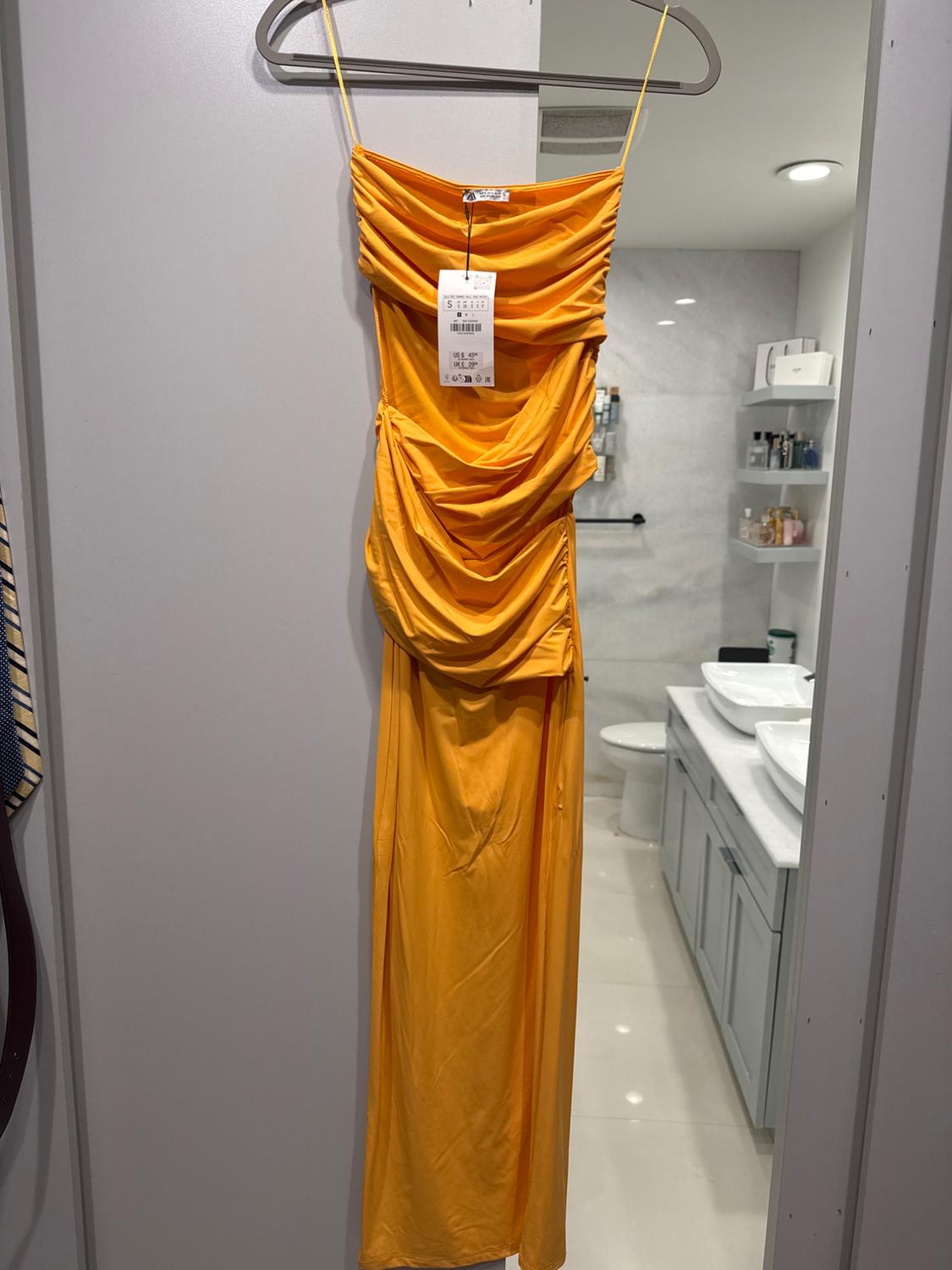 Long yellow dress with thin straps and draped, hanging on a hanger in a modern bathroom. Visible tag in the center.