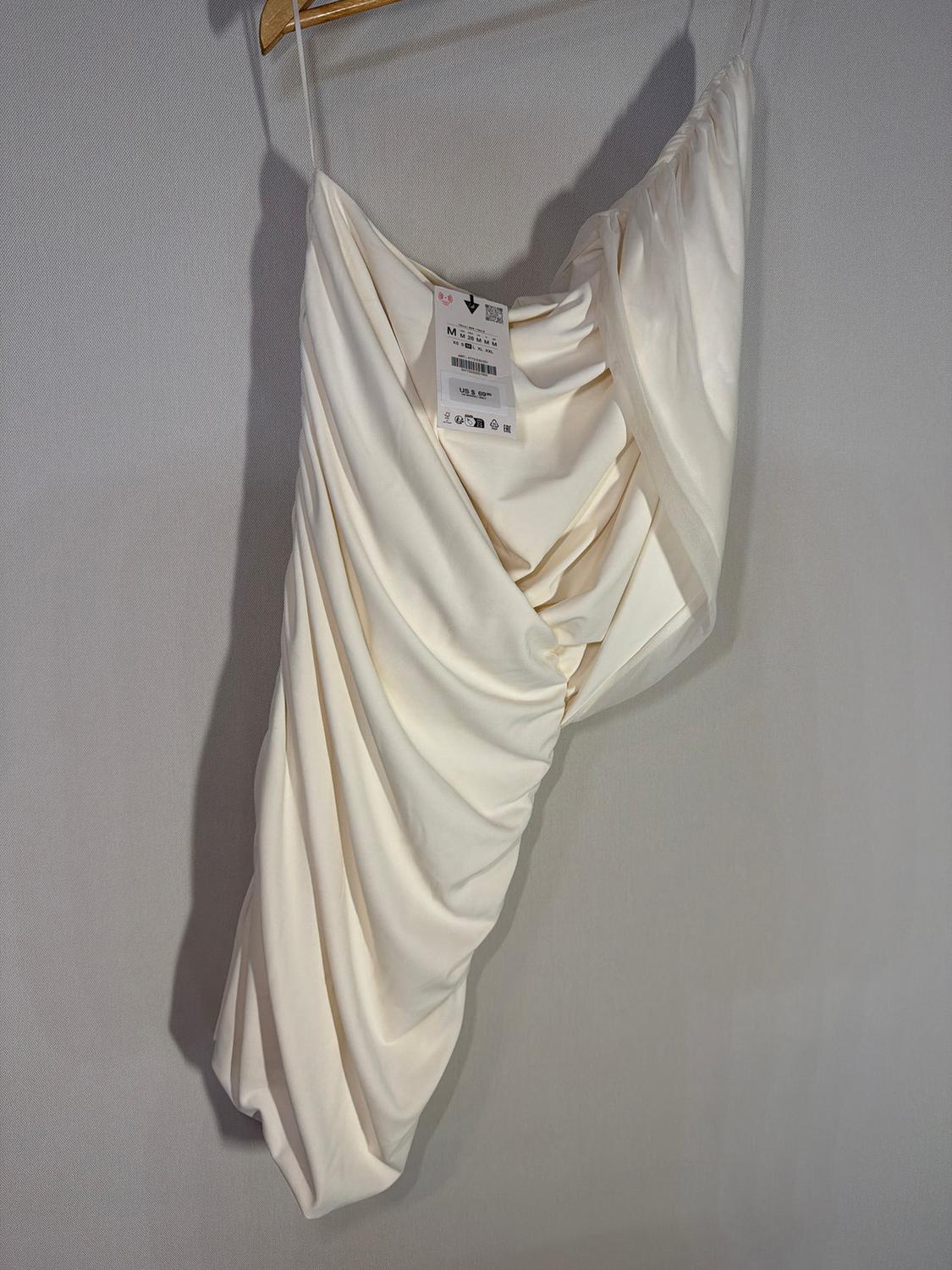 White draped dress hanging on a wooden hanger, with a visible tag showing product details. Light grey background.