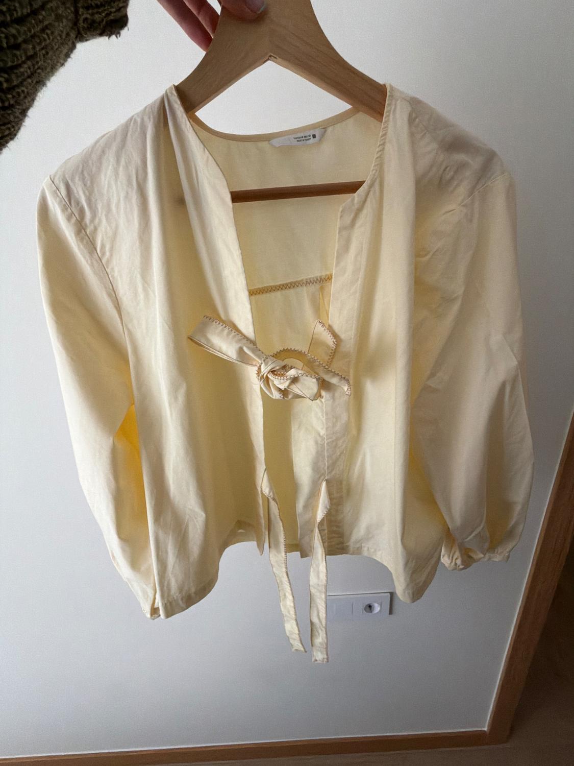 Yellow long-sleeved blouse with a bow on the front, hanging on a wooden hanger, white background.
