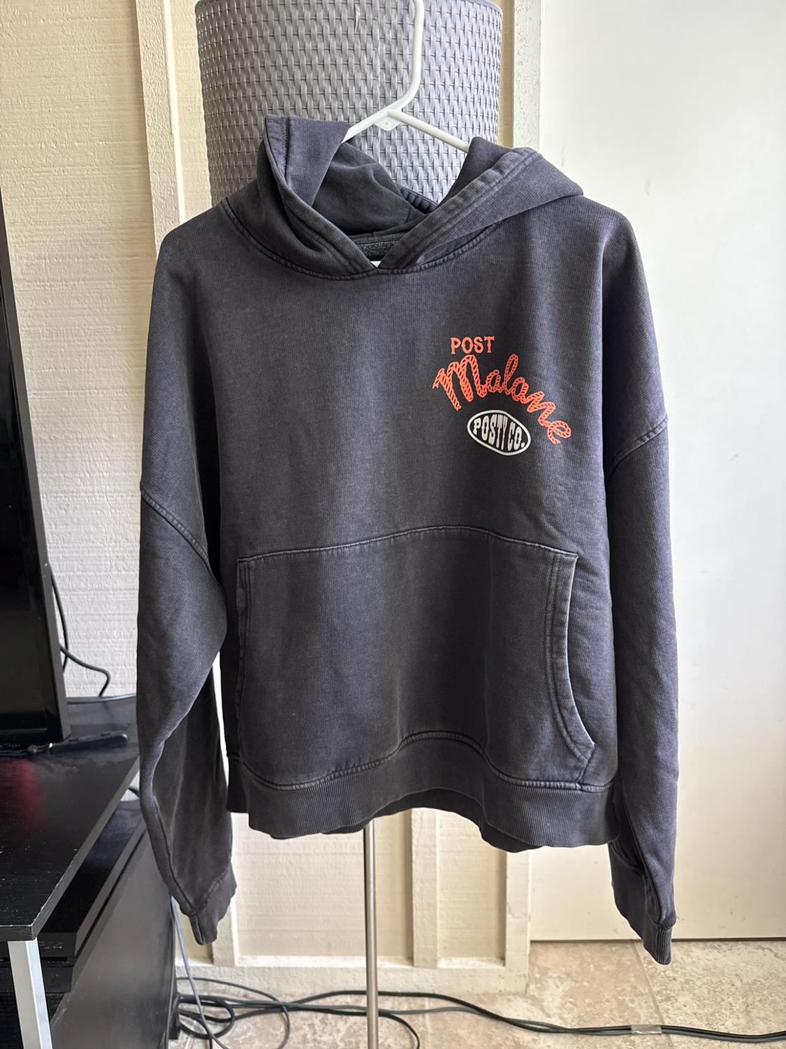 Dark grey hooded sweatshirt hanging in an indoor setting. A front pocket and the text “POST Malone POSTY CO.” on the chest stand out.