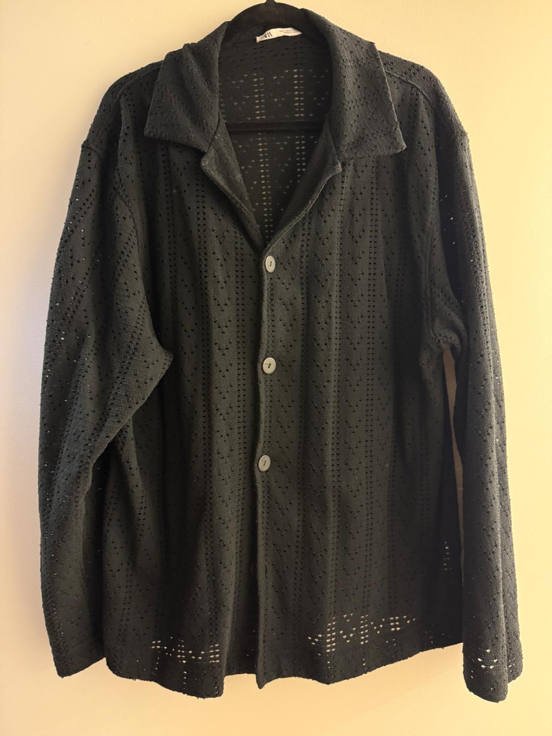 Black lace shirt with white buttons, hanging on a hanger against a beige background. Perforated pattern design.