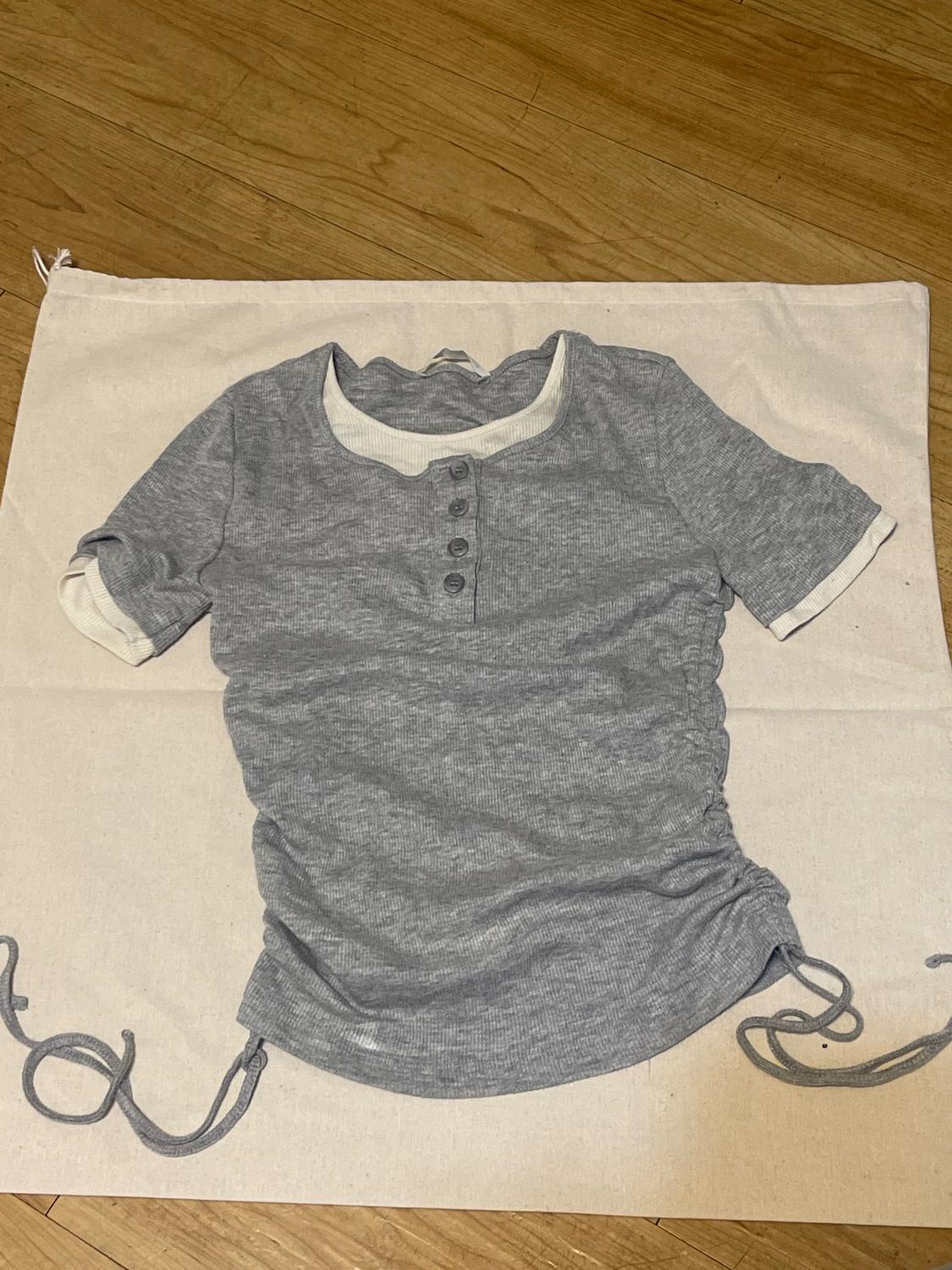 Gray short-sleeved button-up shirt with side laces, on white fabric on a wooden floor.