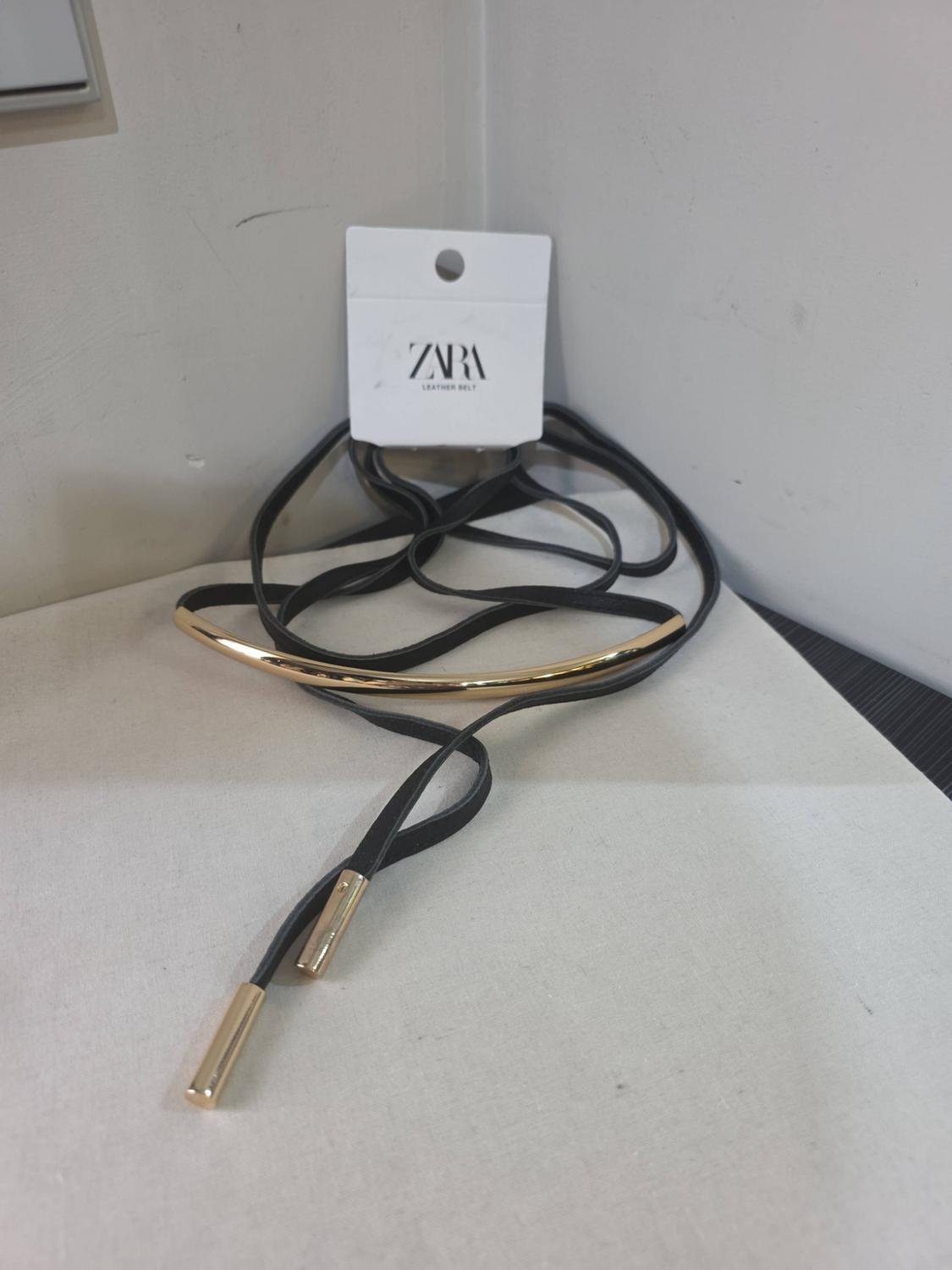 Black belt with a gold-tone metal detail and laces on a light-coloured surface. Label with the text ZARA LEATHER BELT.