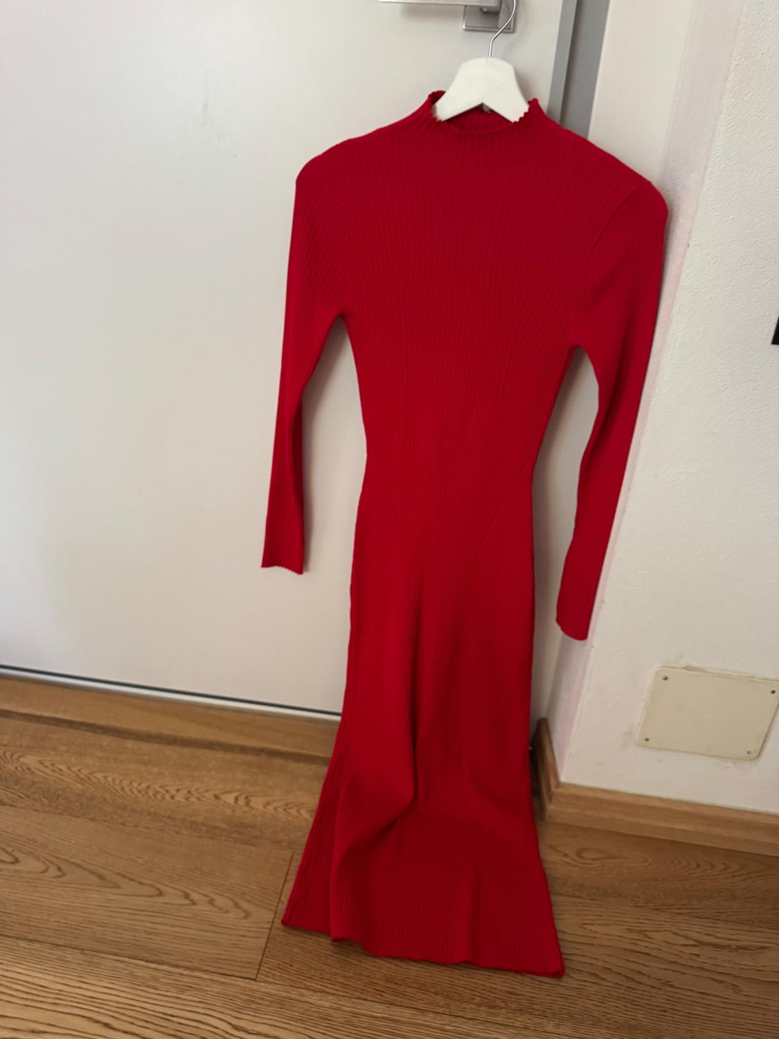 Red knitted dress with long sleeves and a high neck, hanging on a white hanger against a background of white wall and wooden floor.