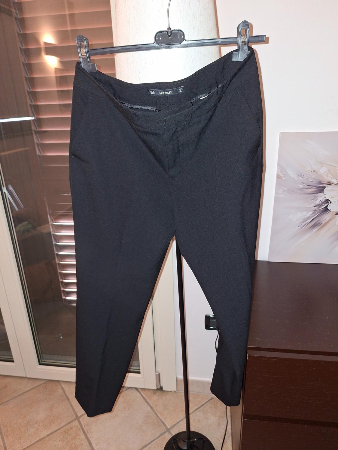 Black trousers hanging on a hanger, with a "Zara Basic" label, in an interior with warm lighting and a background of blinds and furniture.