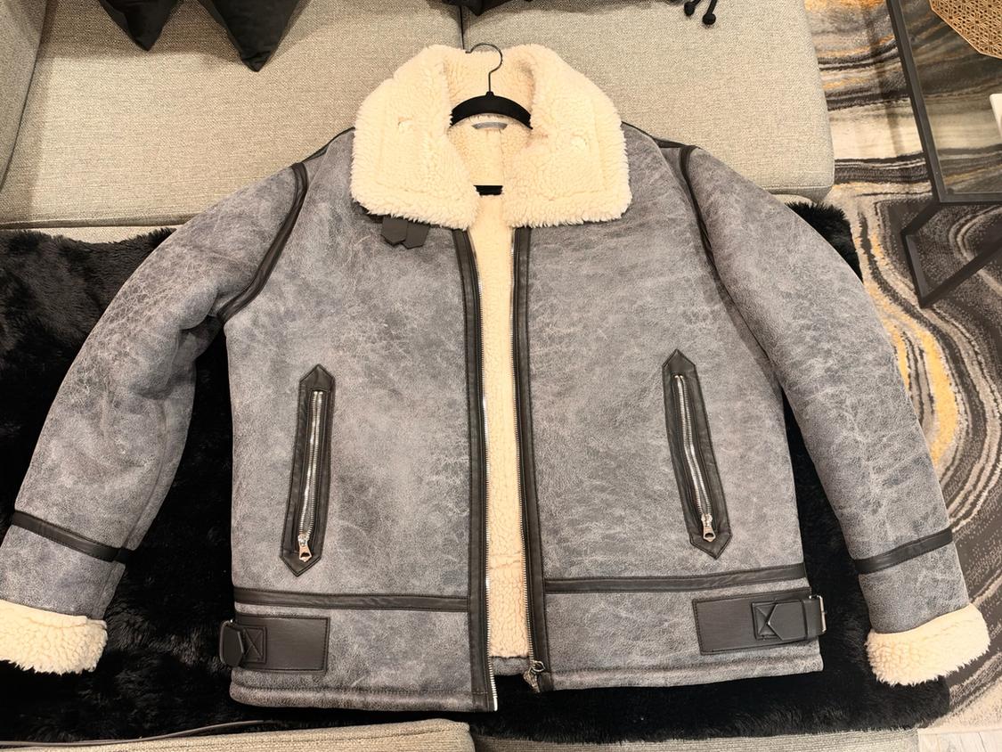 Gray aviator-style jacket with a cream fur collar and lining, black details, zippered pockets and buckles, hanging over a sofa.