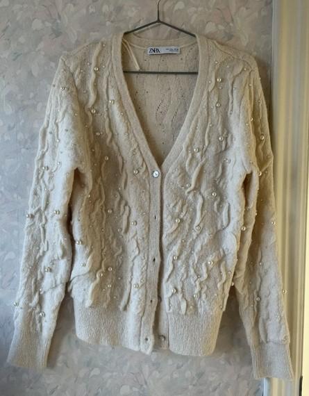 Beige textured cardigan with decorative pearls, hanging on a hanger. Light wall background with floral design. Label visible.