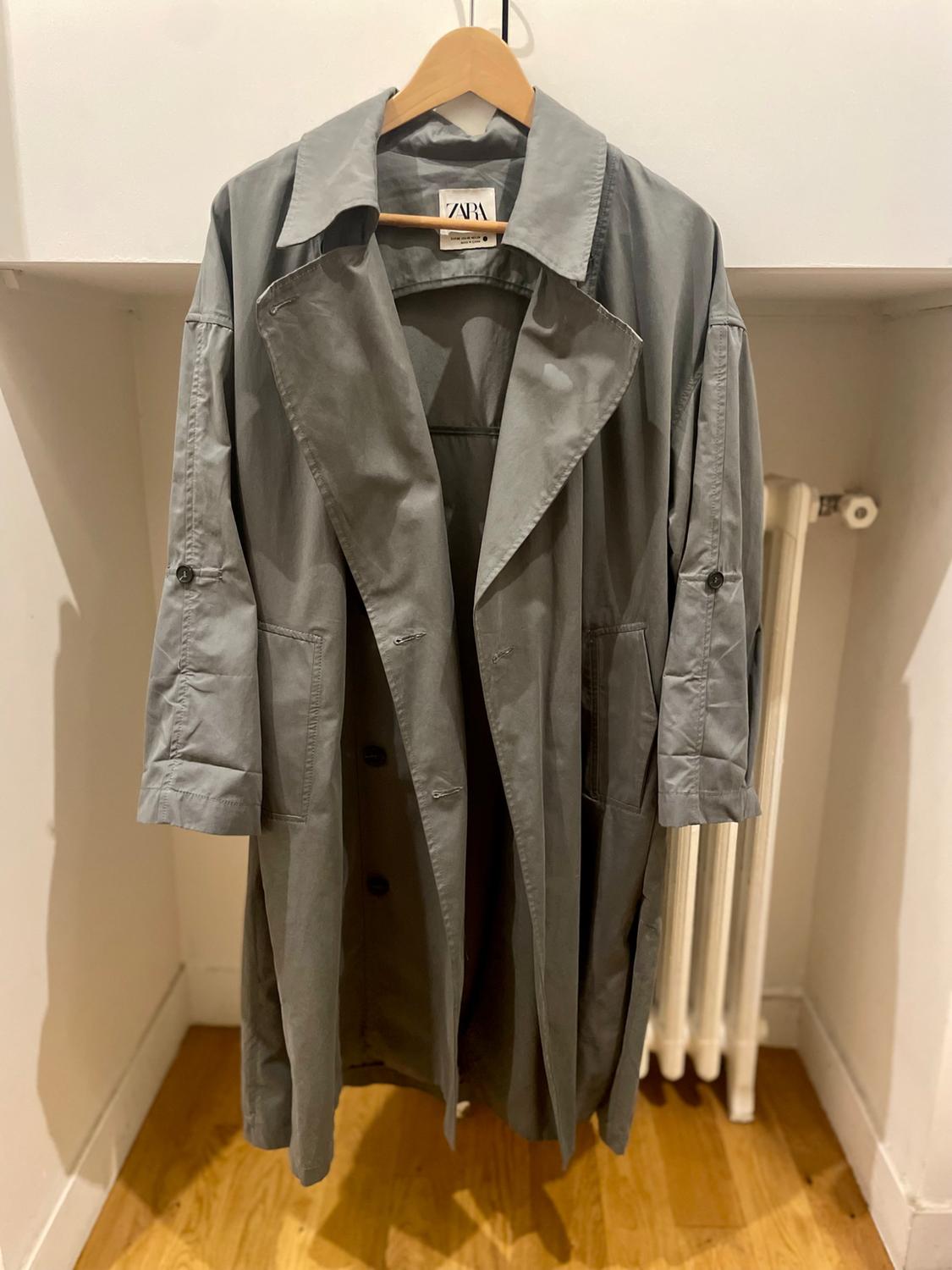 Grey trench coat hanging on a wooden hanger, with a visible Zara label. White wall background and light wooden floor.