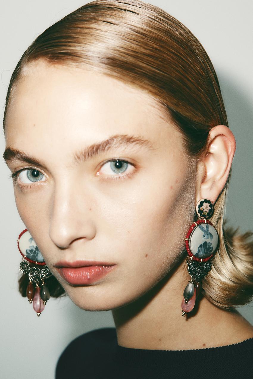 Close-up of gold earrings with oval resin pendants, beads, and contrasting metallic flowers.