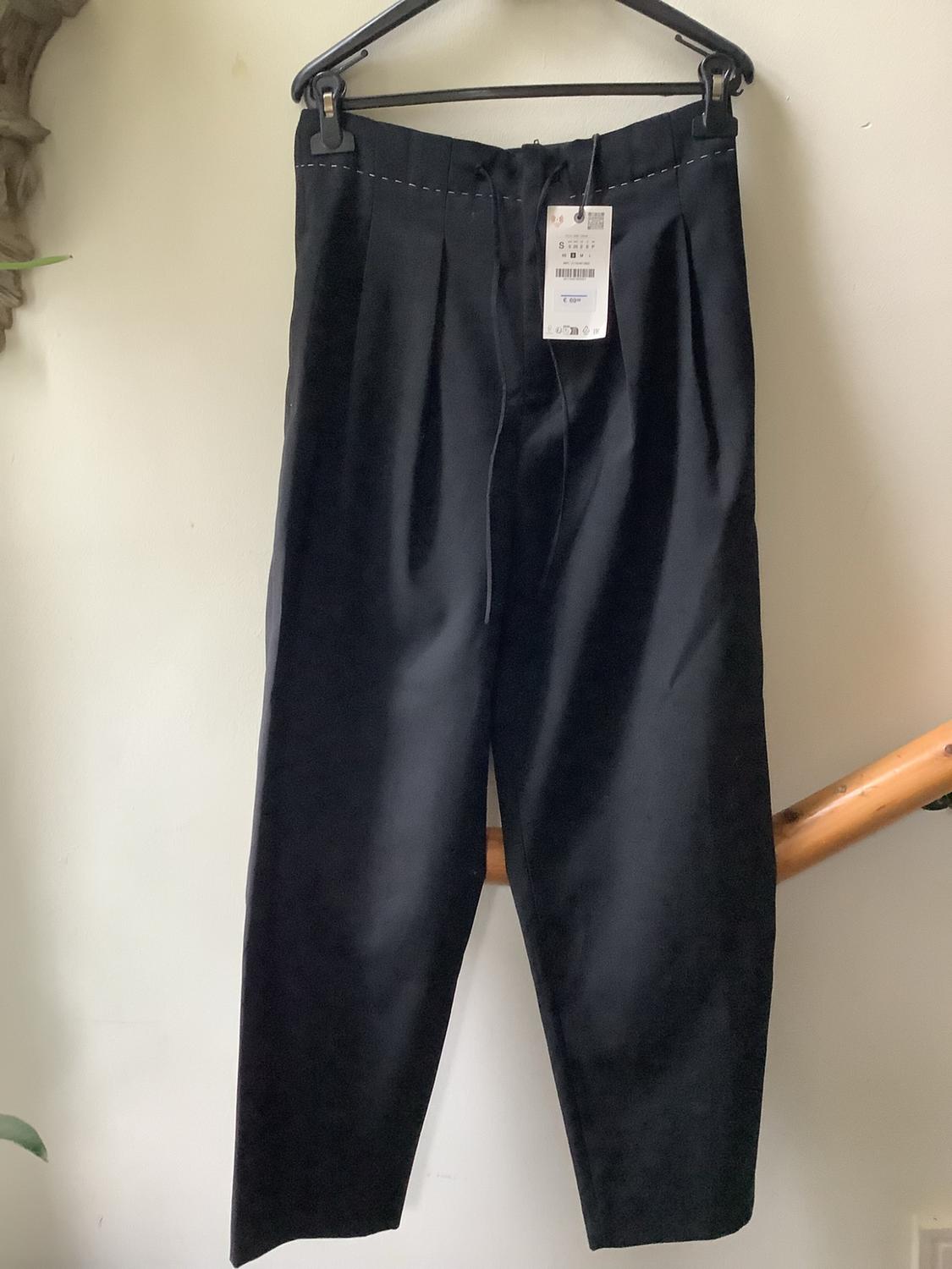 Black pleated trousers with a drawstring and white stitching detail, hanging on a hanger against a white wall next to a handrail.