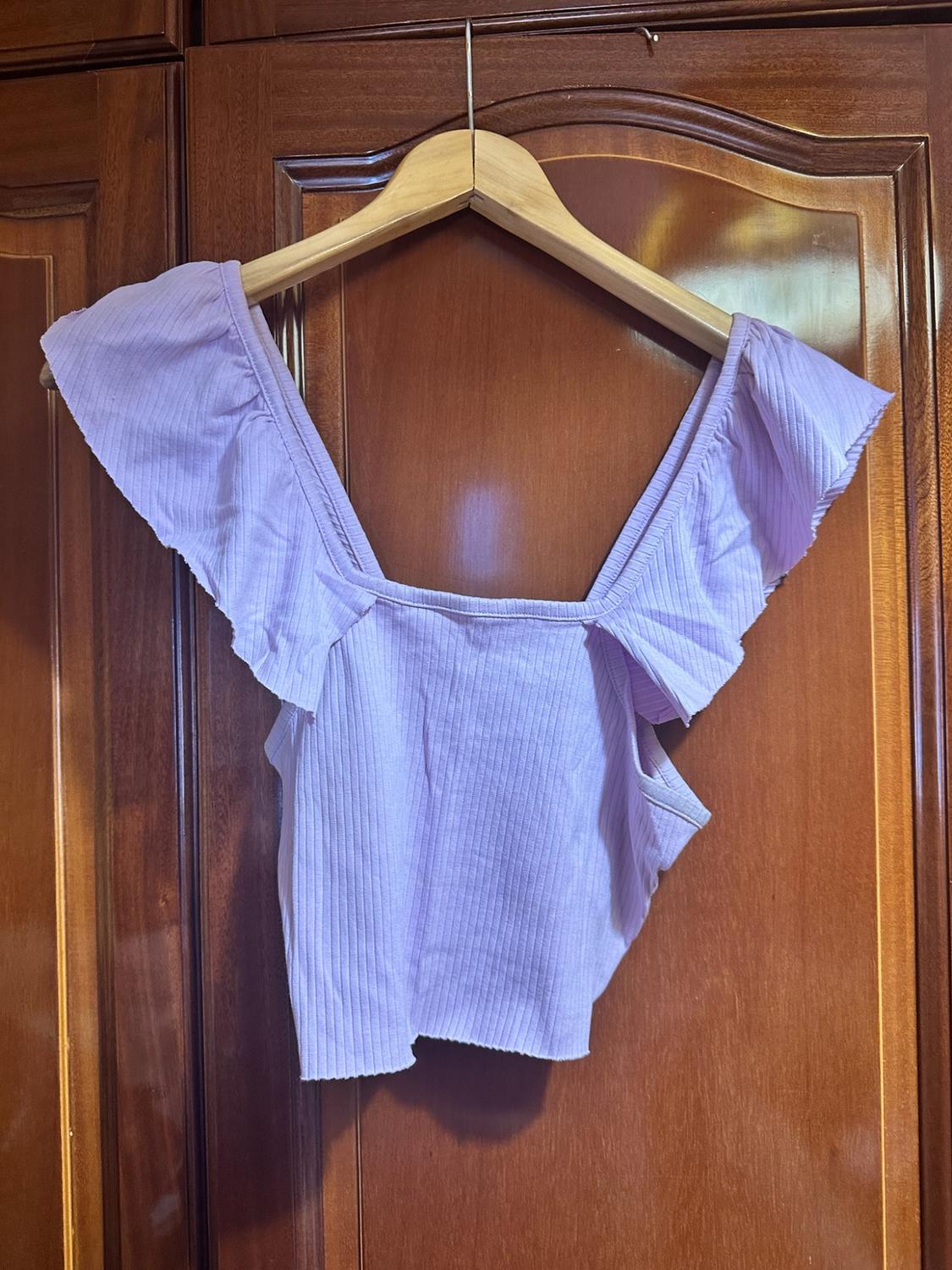 Lilac ribbed crop top with frills, hanging on a hanger in front of a wooden wardrobe.
