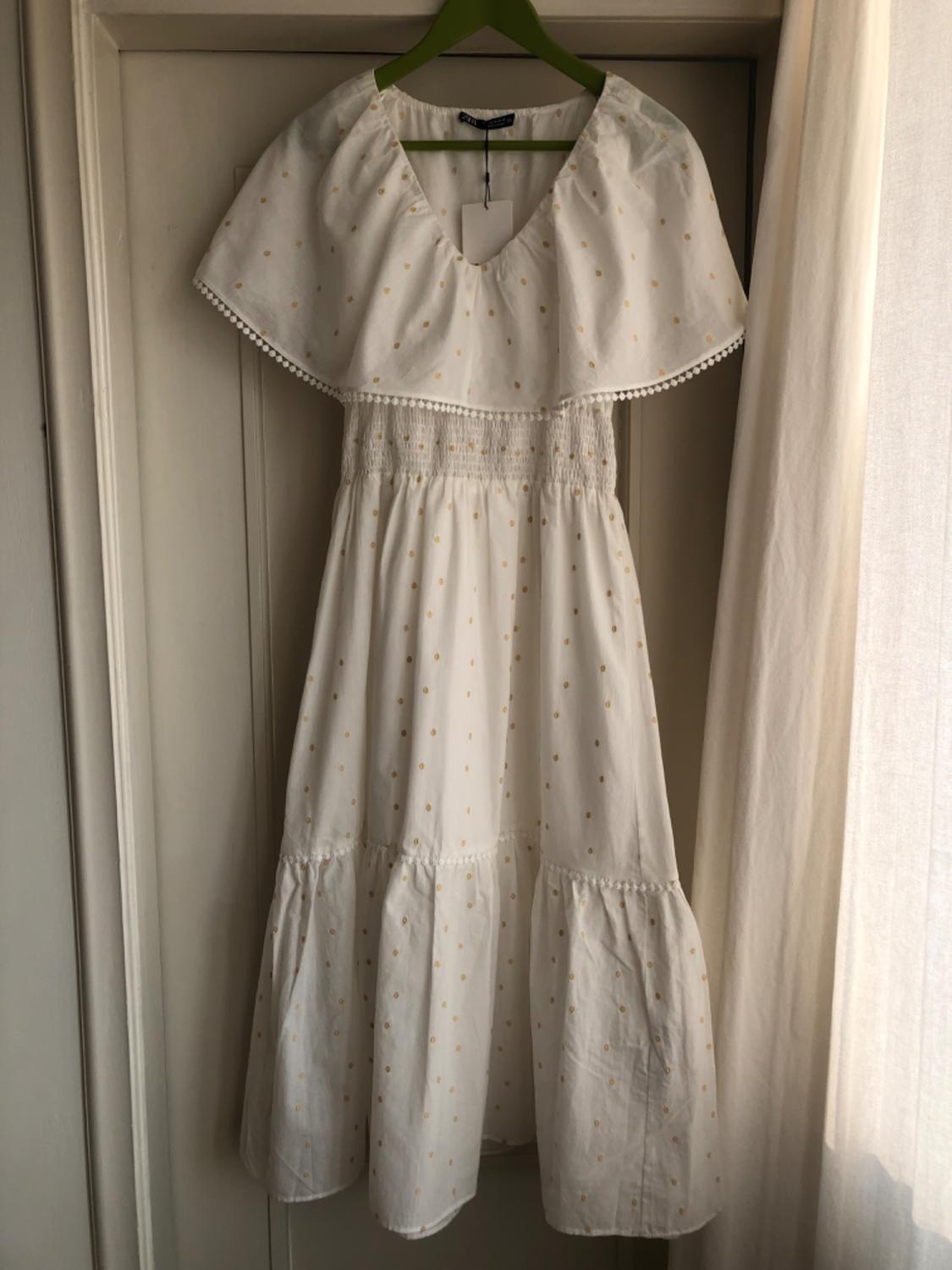 White dress with a pattern of small yellow dots, flared design and lace detailing, hanging on a green coat hanger in front of a curtain.