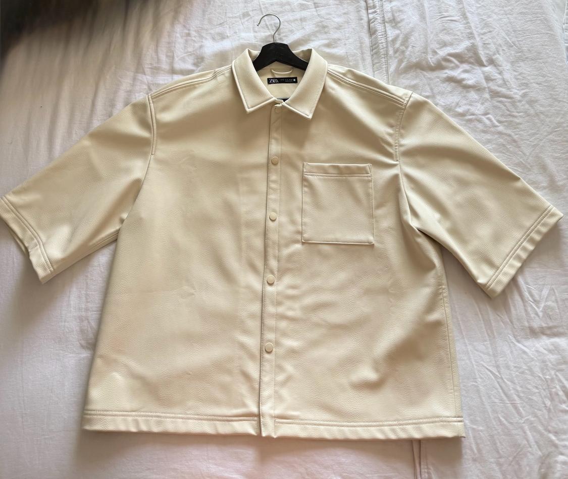 Cream-coloured short-sleeved shirt with press stud buttons and a pocket, on a black hanger on a crumpled white sheet. ZARA label.