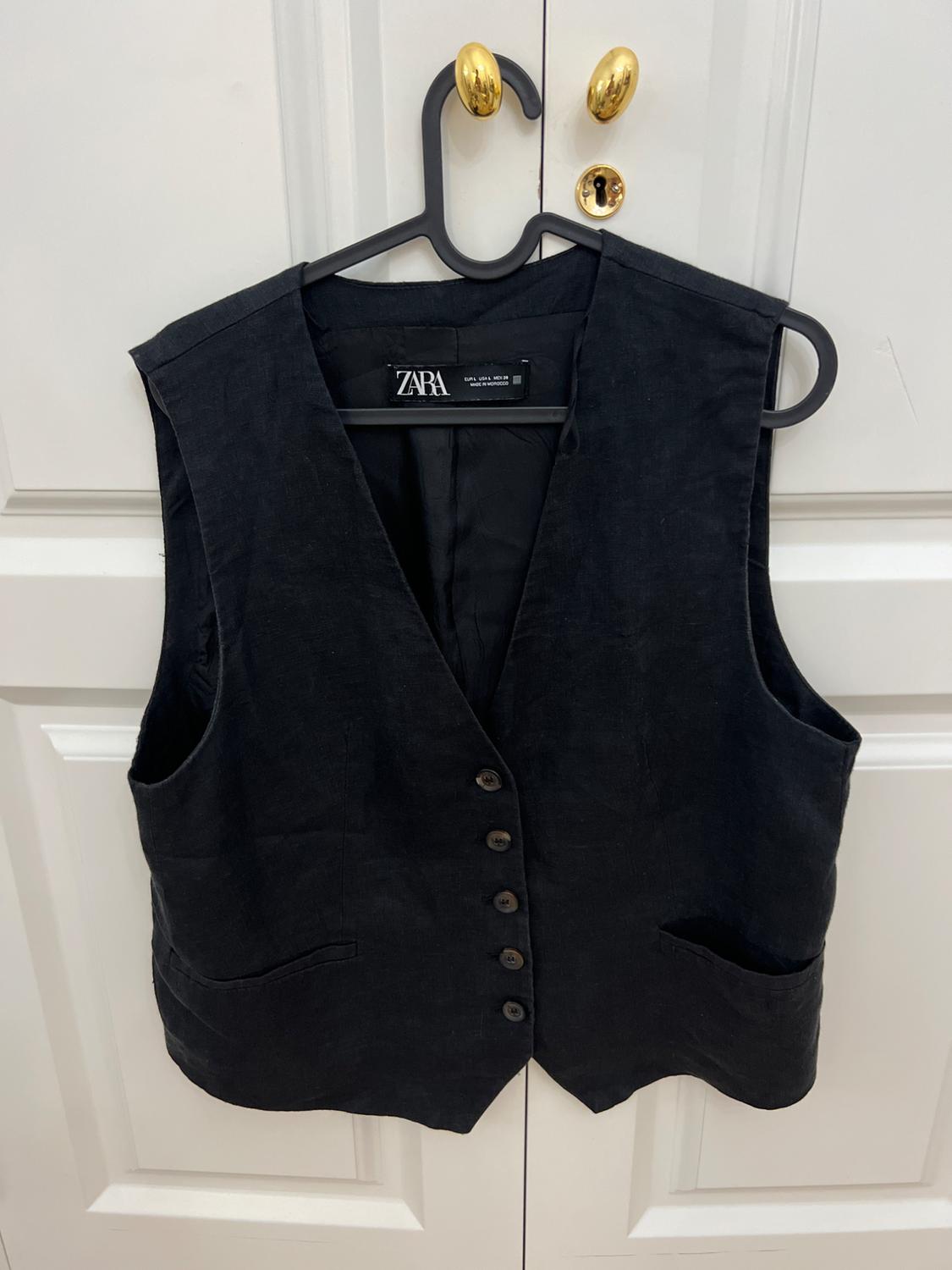 Casual-style black button-up waistcoat hanging on a coat hanger against a white door. Label with the text ZARA.