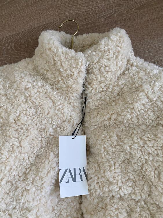 Beige fleece jacket hanging on a golden hanger over a wooden floor. White tag with the text "ZARA".