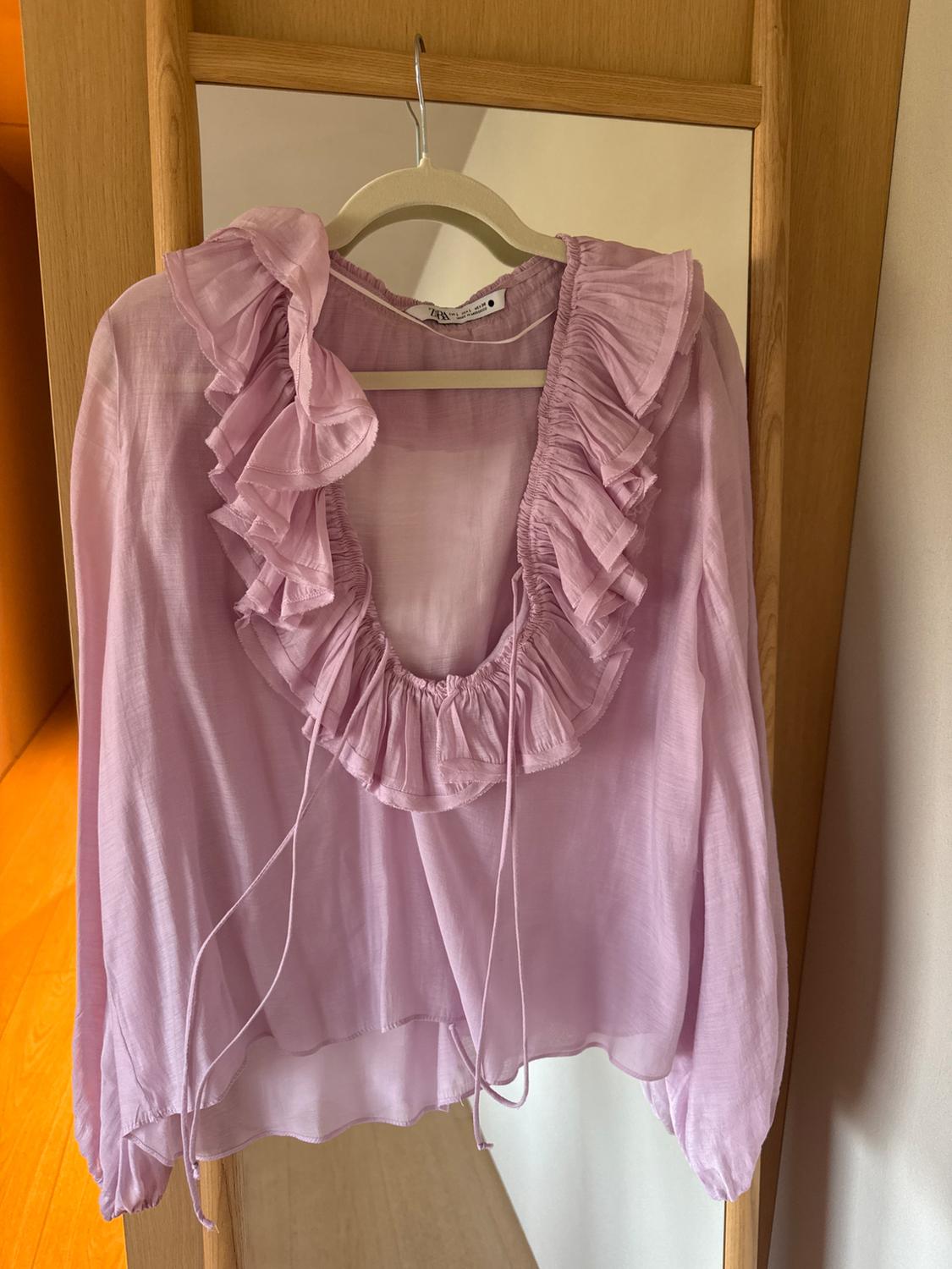 Pink blouse with ruffles on the neckline and long sleeves, hanging on a wooden hanger in front of a mirror.