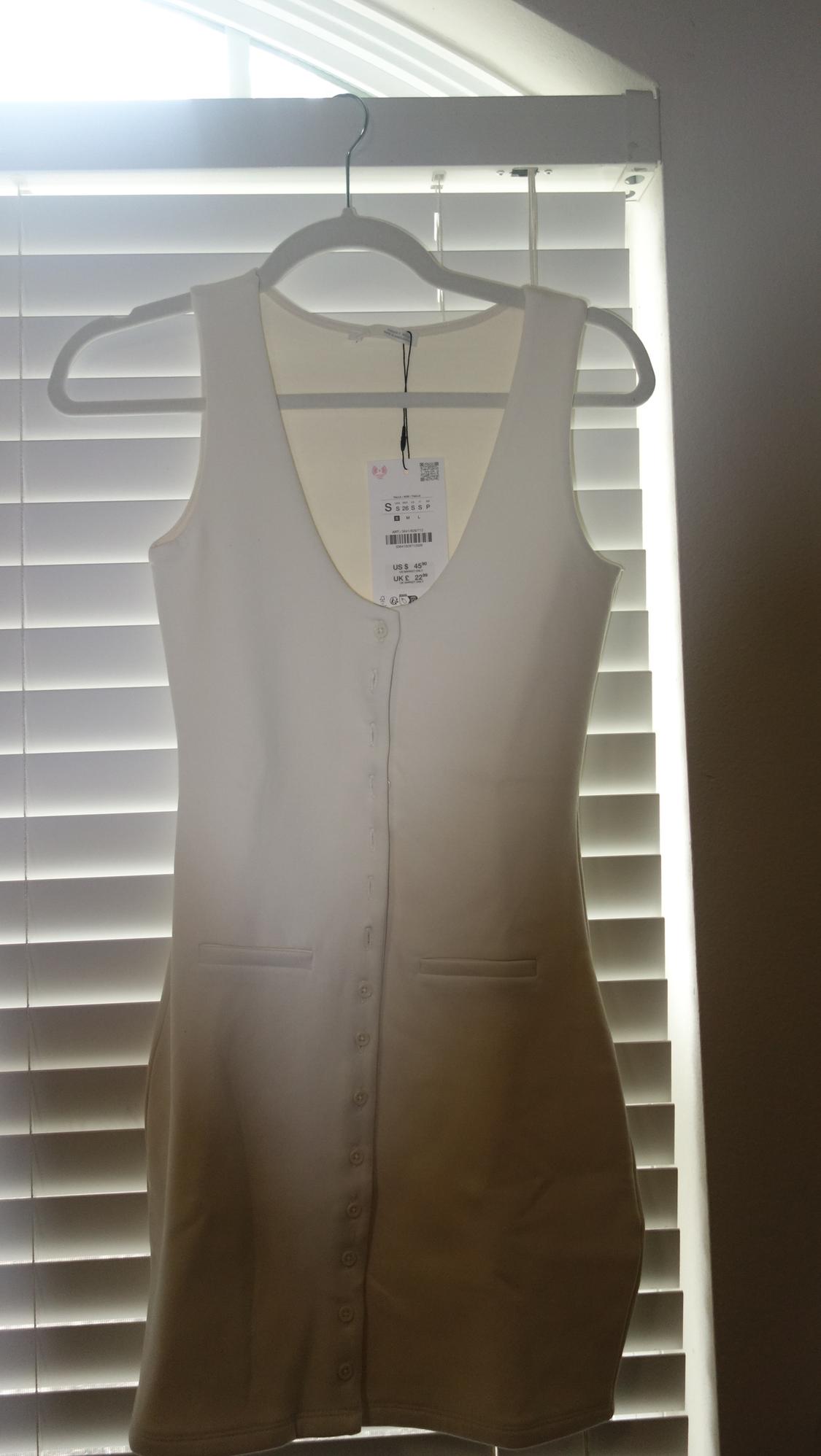 Sleeveless white dress hanging on a hanger in front of a window with blinds. Visible tag with size and price details.