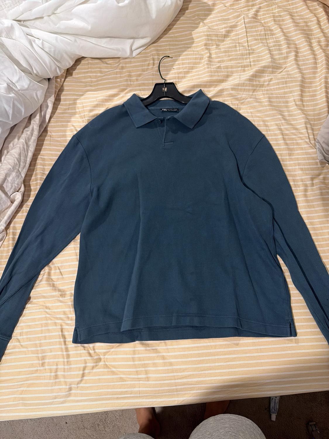 Long-sleeve polo-style shirt in blue on a black hanger, placed on a bed with beige and white striped sheets.