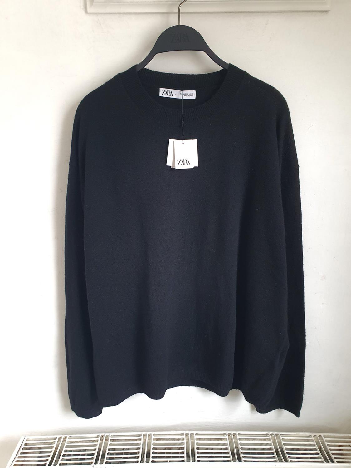 Black jumper hanging on a black hanger with a visible Zara label. White background and metal grid at the bottom.
