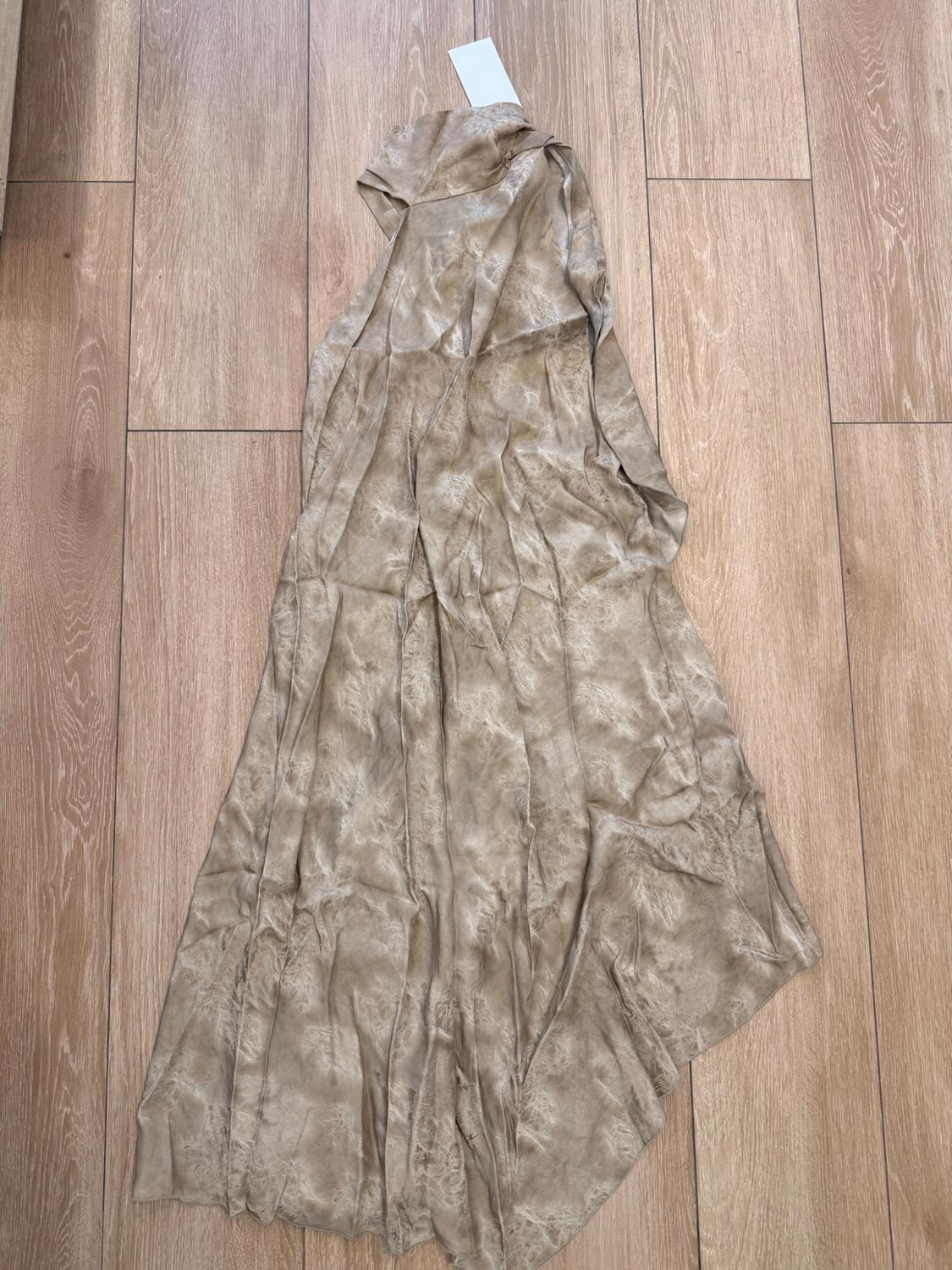 Long garment in beige satin fabric with a subtle pattern, laid out on a light wood floor. Includes a white label.