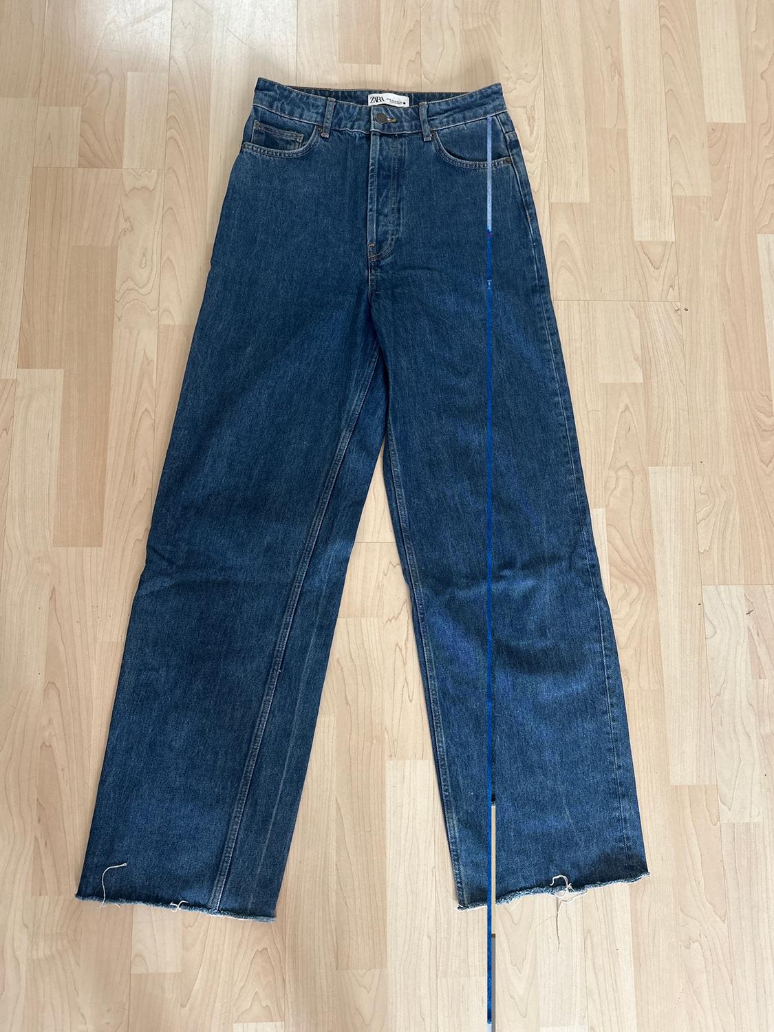 Wide-leg blue denim jeans with a frayed hem, laid out on a light wooden floor. Visible waist label.