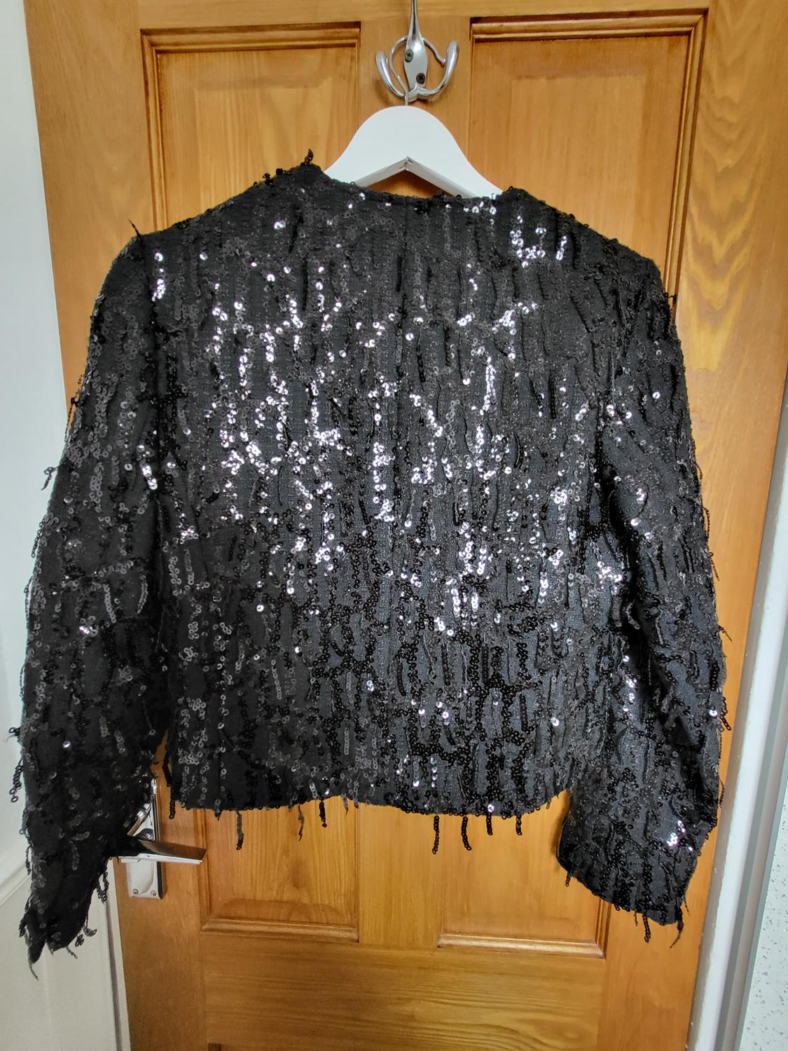Black sequinned jacket with a fringed hem, hanging on a white hanger against a wooden door.